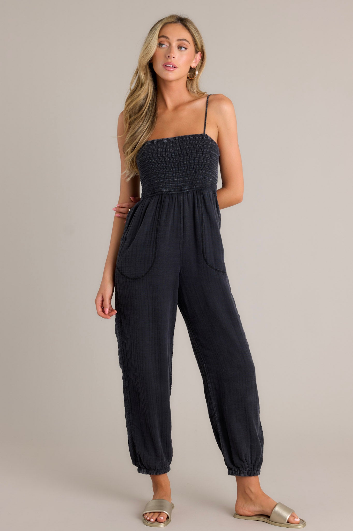 Front view of a black jumpsuit featuring a square neckline, thin adjustable straps, a fully smocked bodice, functional hip pockets, a wide leg design, and elastic cuffed ankles.