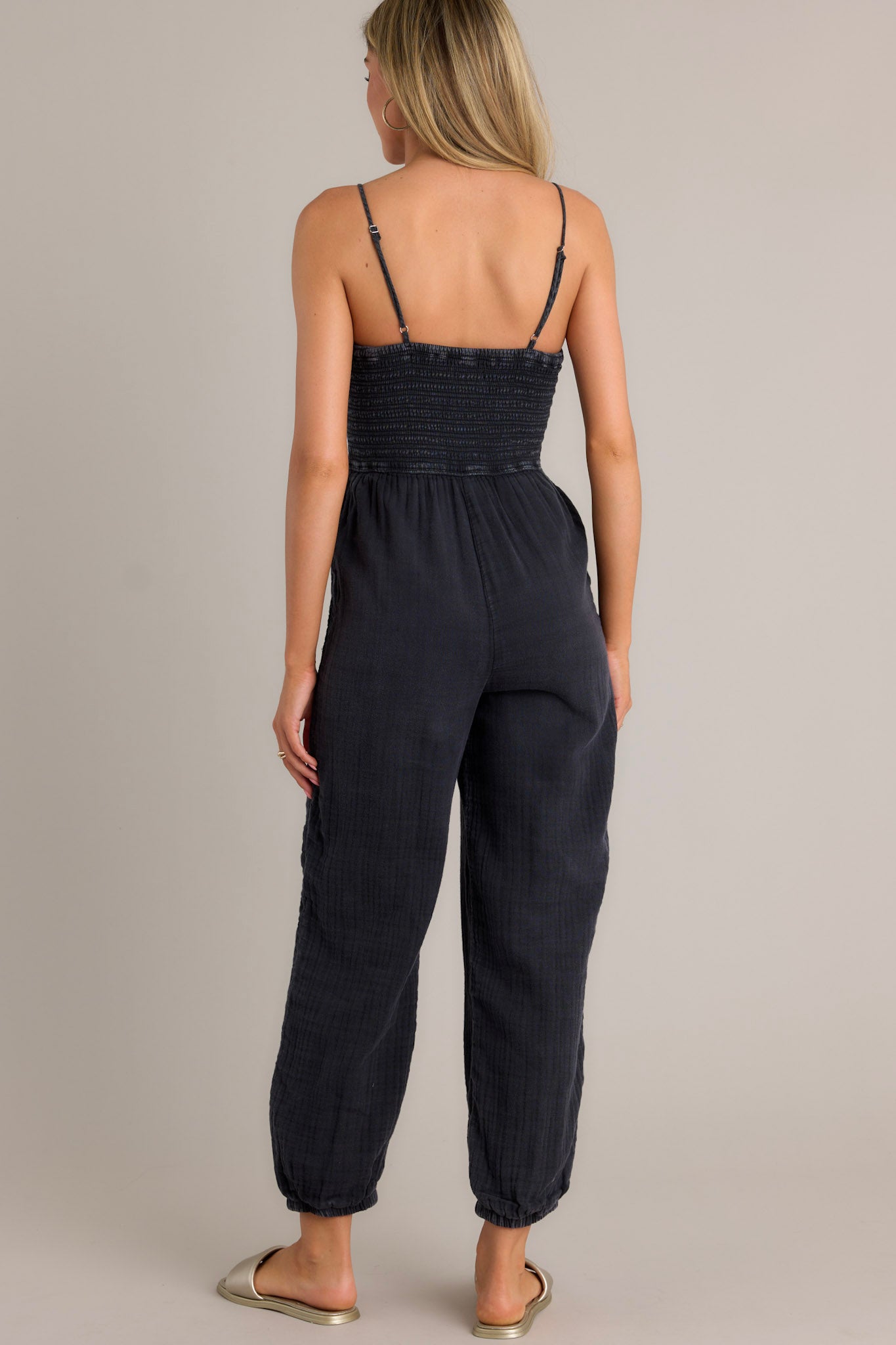 Back view of a black jumpsuit highlighting the thin adjustable straps, fully smocked bodice, and overall fit.