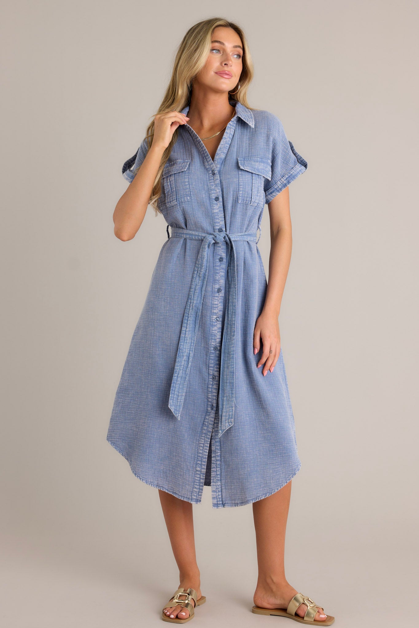 Front angled view of a blue maxi dress featuring a collared v-neckline, a functional button front, functional breast pockets, a self-tie waist belt, belt loops, and cuffed short sleeves