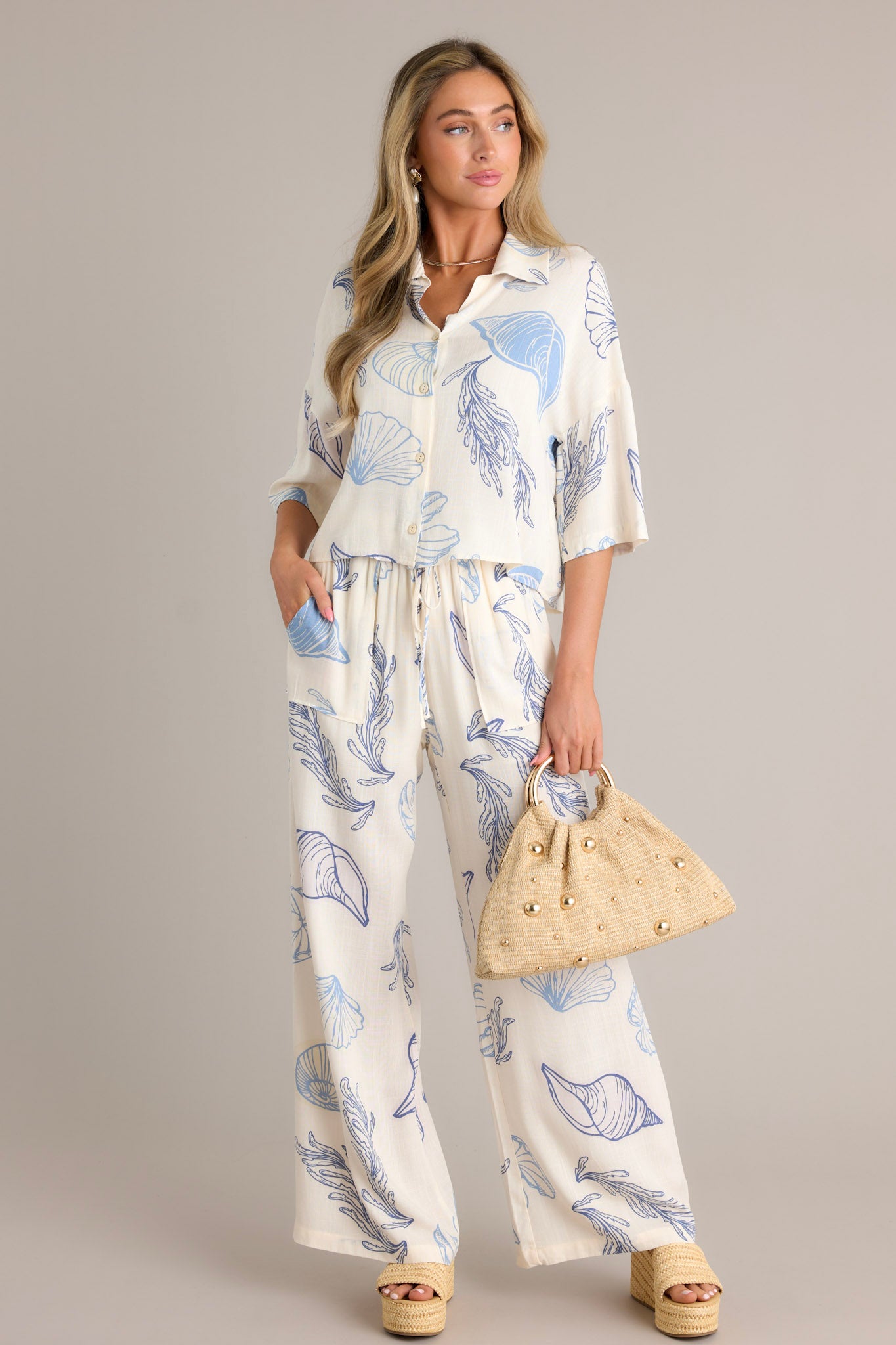 Front angled view of pants featuring an elastic waistband, a drawstring tie, a wide leg design, and a beachy seashell print throughout