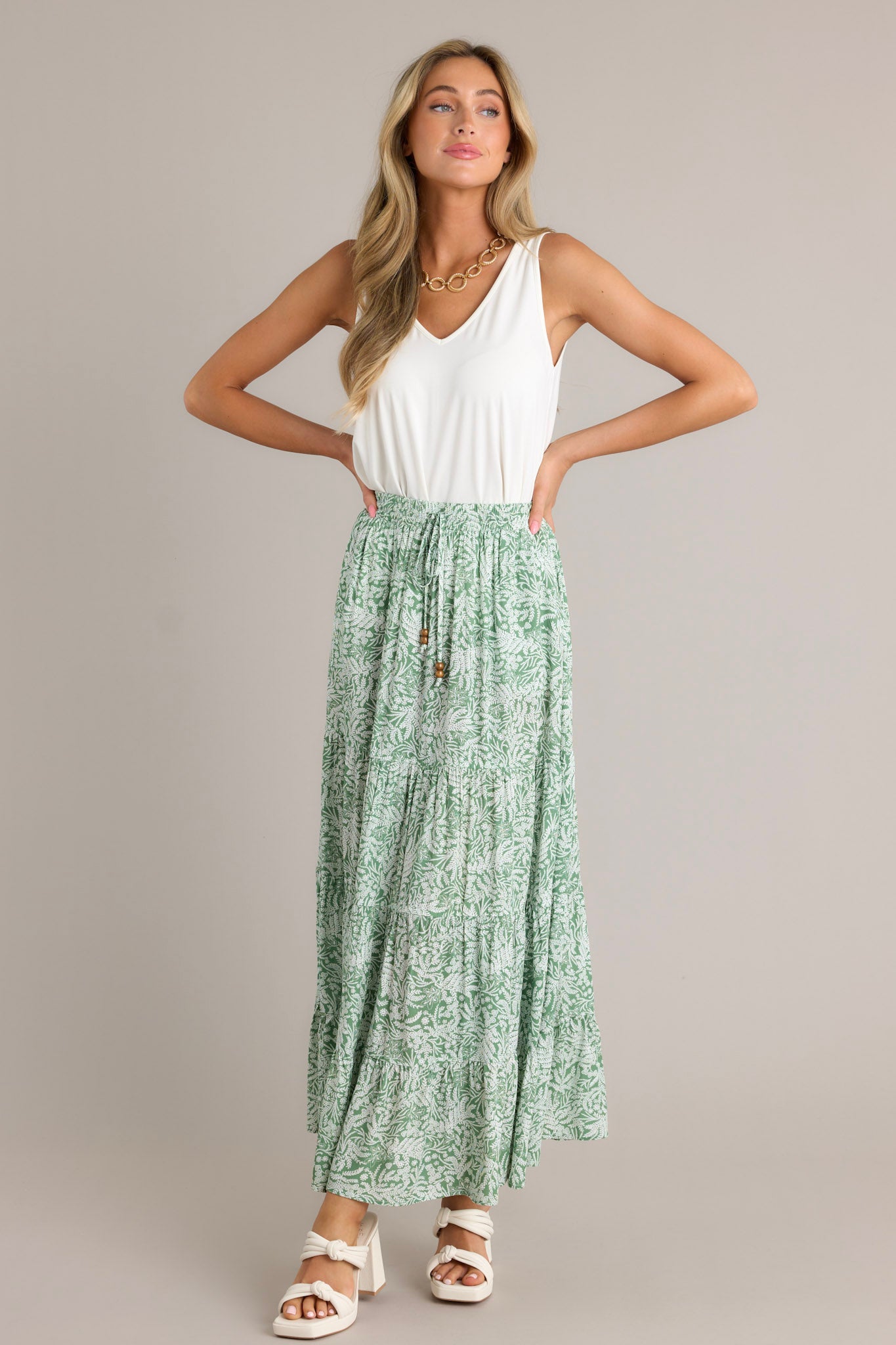 Full length view of a green maxi skirt with a high waisted design, an elastic waistband, a self-tie drawstring, multi tiers, and a flowing silhouette