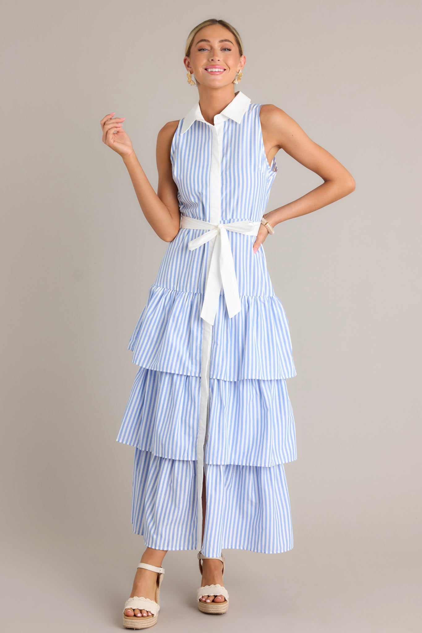This blue stripe maxi dress features a collared neckline, a button front, an elastic waist insert, a self-tie waist belt, and multiple tiers.