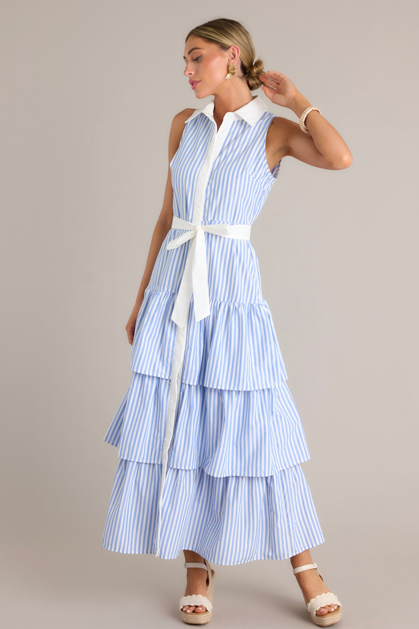 Front angled view of a blue stripe maxi dress featuring a collared neckline, a button front, an elastic waist insert, a self-tie waist belt, and multiple tiers