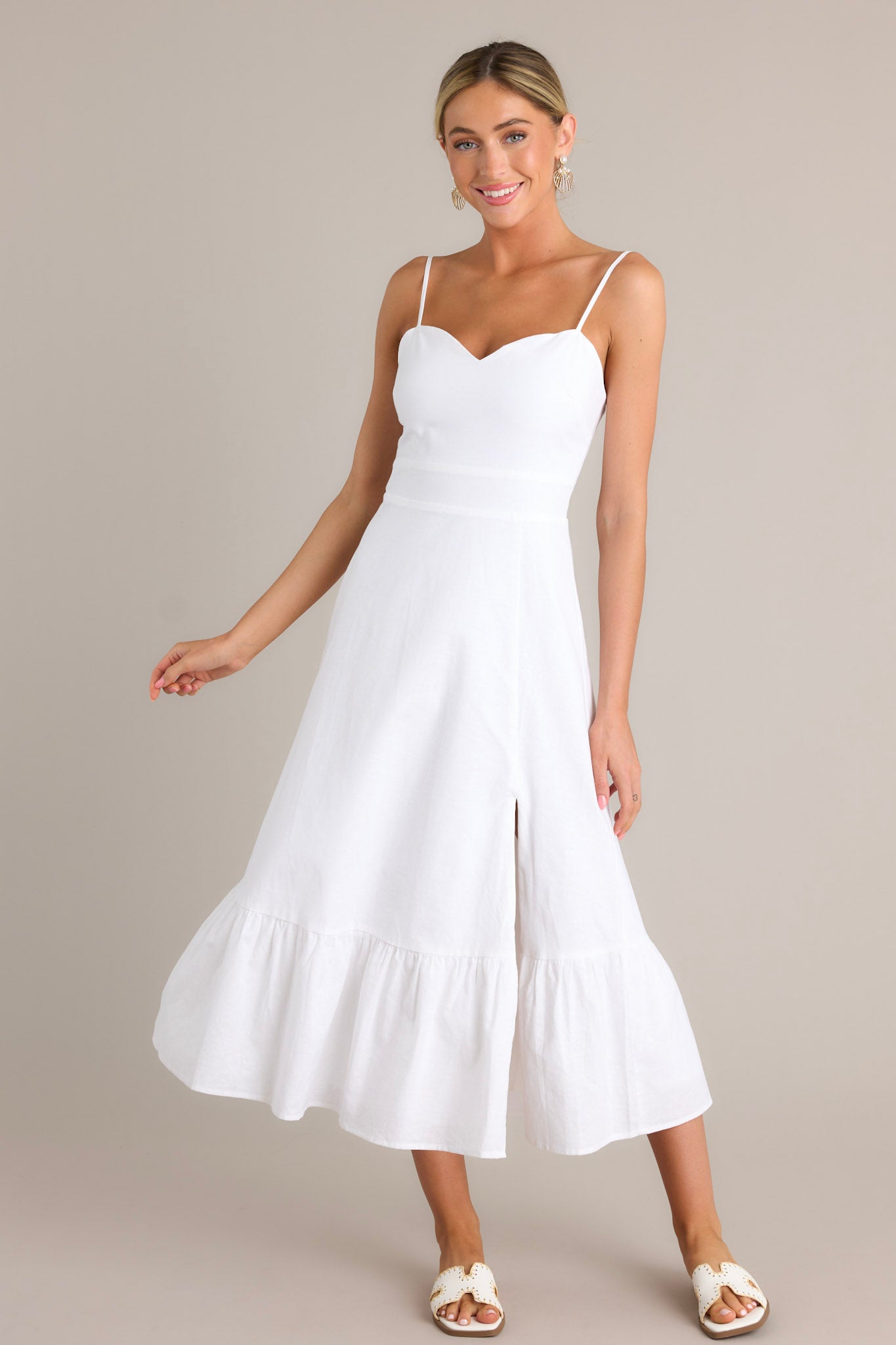 Front angled view of a white maxi dress featuring a sweetheart neckline, thin adjustable straps, a smocked back insert, a discrete zipper, a single tier, and a slit