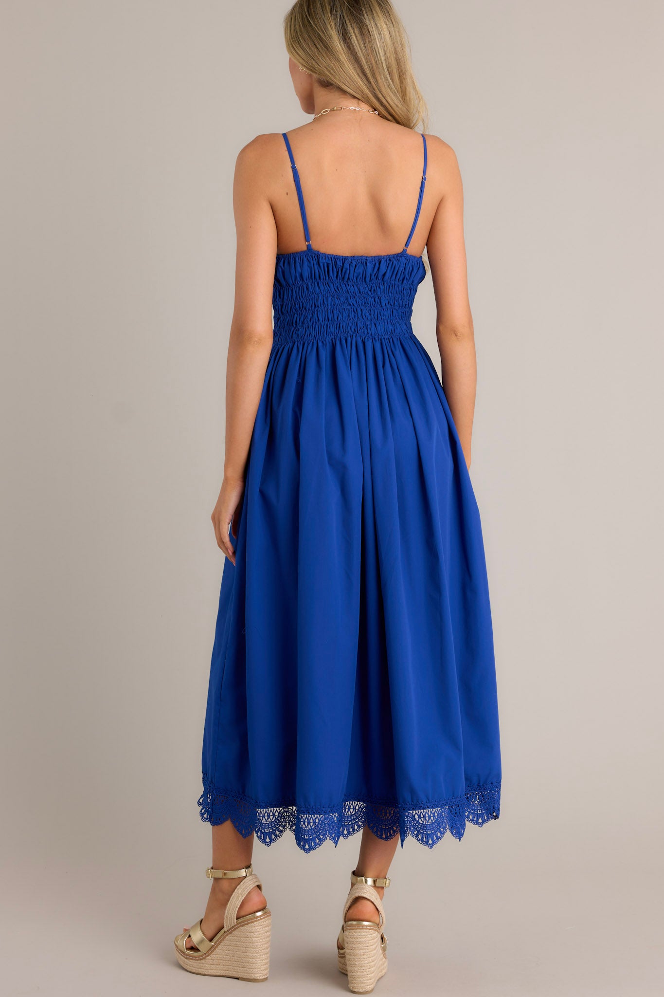 Back view of a blue maxi dress highlighting the fully smocked waist & back, thin adjustable straps, and lace hemline.