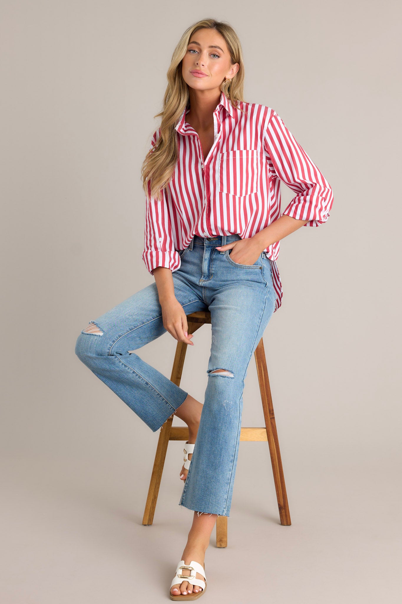 Seated full length view of a red stripe top with a collared neckline, a functional button front, a breast pocket, rollable button cuffed sleeves, and a split high-low hemline