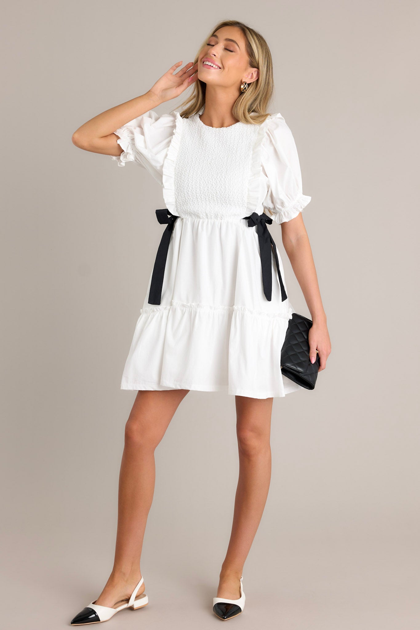 Front angled view of an ivory mini dress featuring a smocked bodice design, self-tie waist bows, a tiered design, a discrete back zipper, and cuffed puffer sleeves