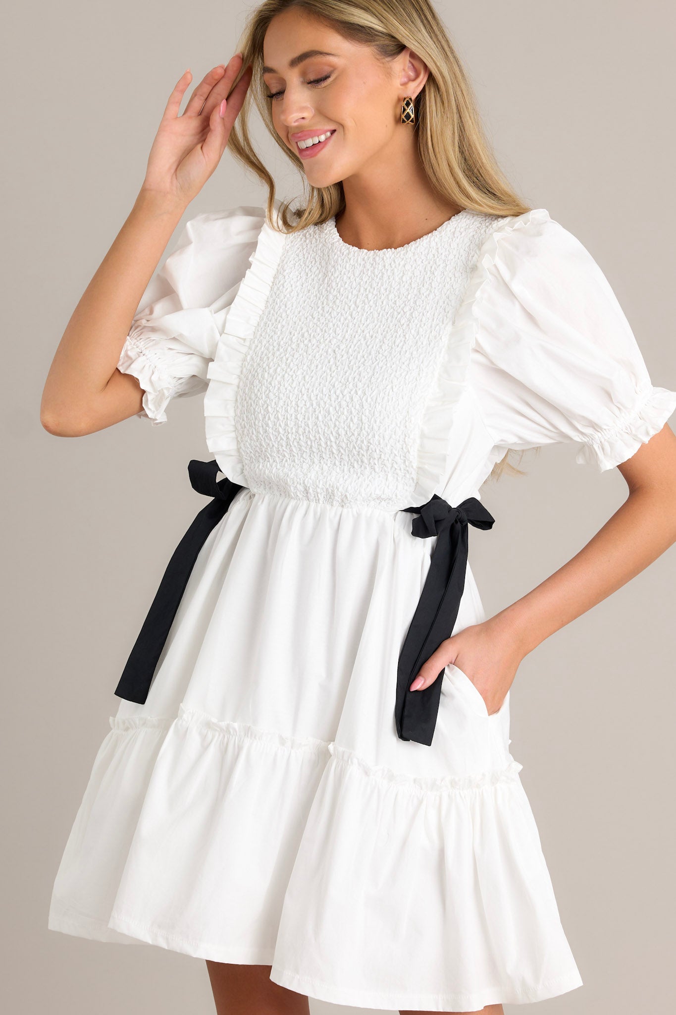 Side view of an ivory mini dress showcasing the smocked bodice design, self-tie waist bows, tiered design, discrete back zipper, and cuffed puffer sleeves.