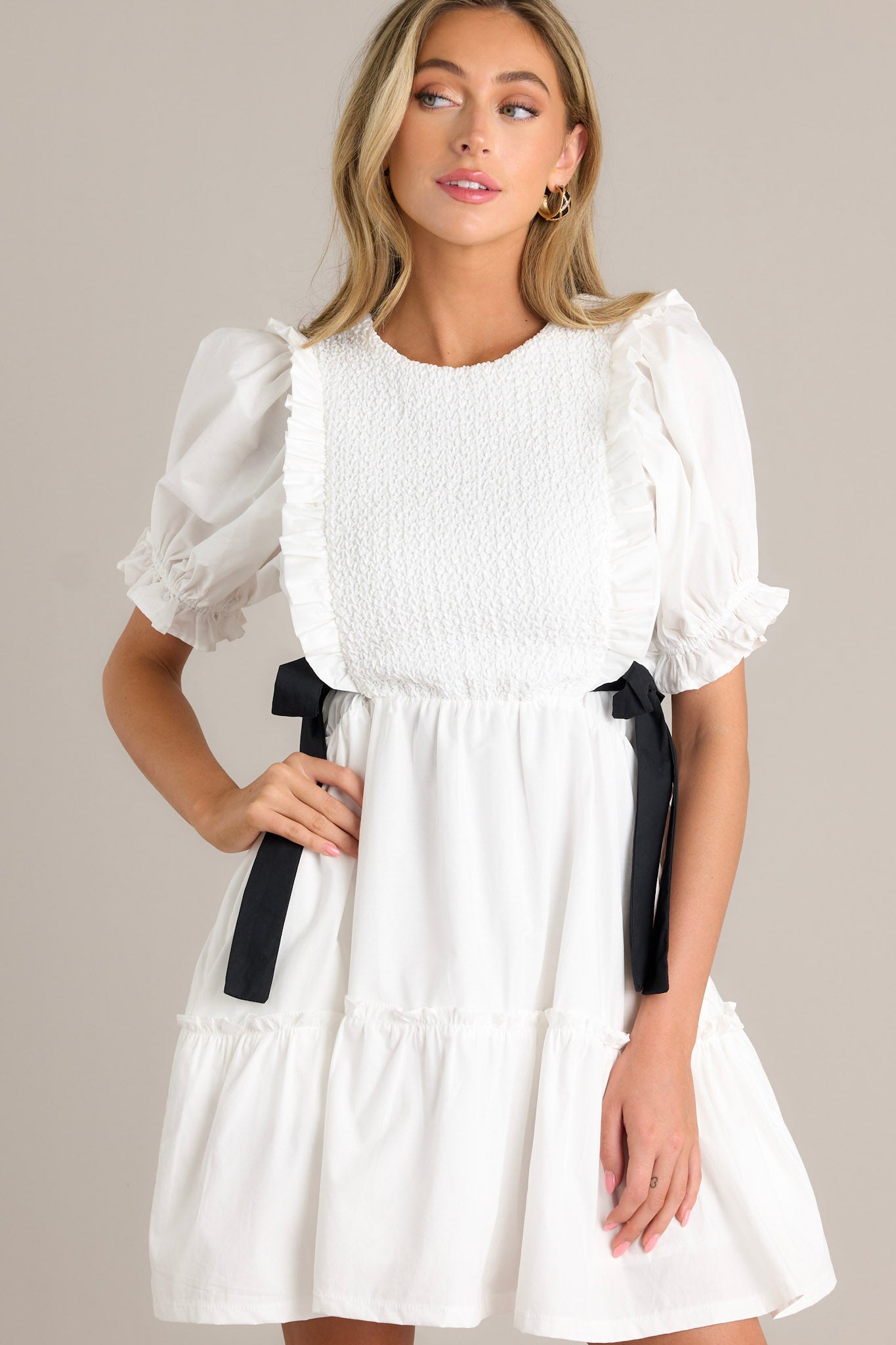 Front view of an ivory mini dress featuring a smocked bodice design, self-tie waist bows, a tiered design, a discrete back zipper, and cuffed puffer sleeves.