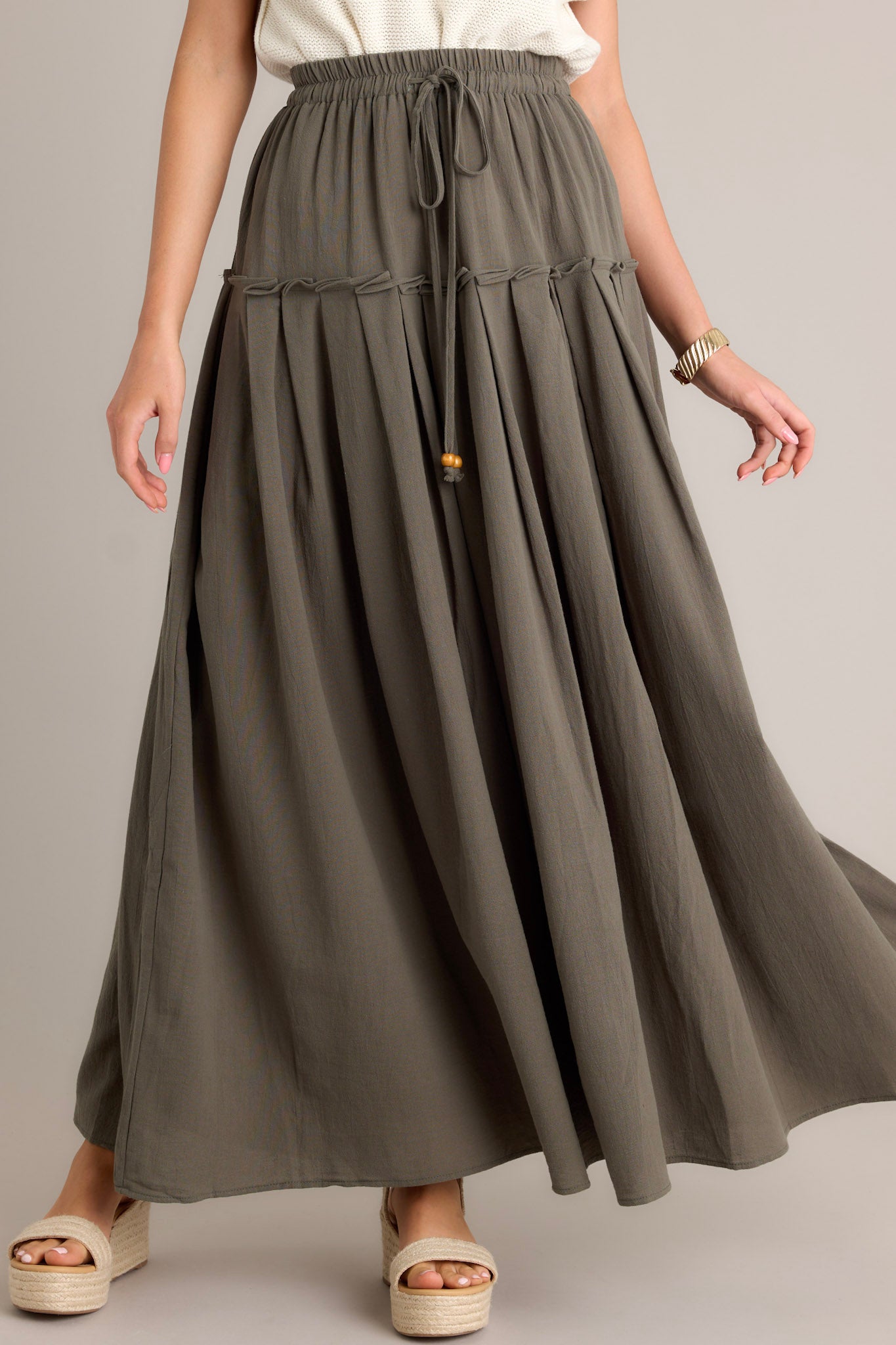 Front view of an olive maxi skirt featuring a high waisted design, an elastic waistband, a self-tie drawstring, a single tier, subtle pleats, and a flowing silhouette.