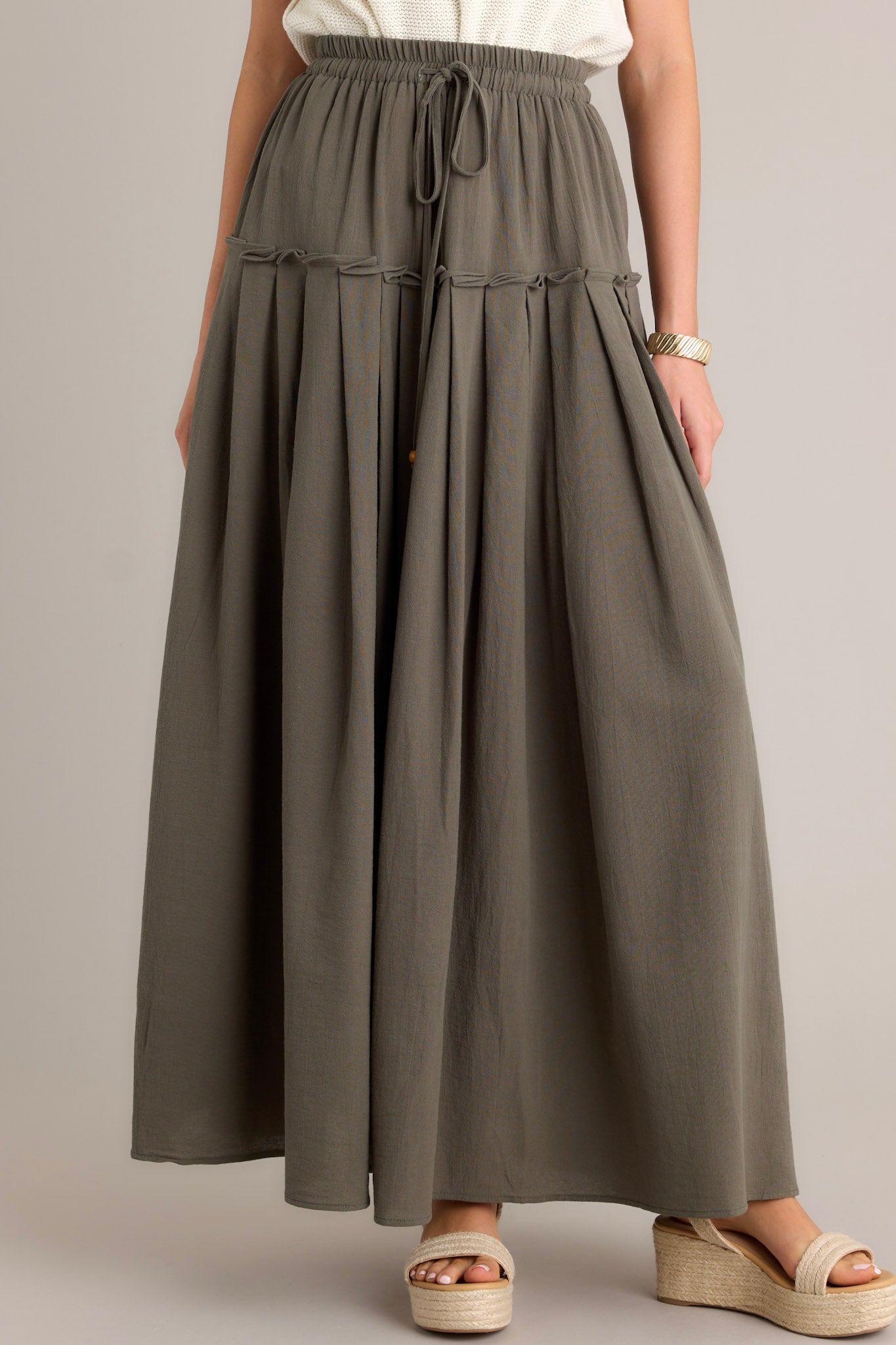 Front angled view of an olive maxi skirt featuring a high waisted design, an elastic waistband, a self-tie drawstring, a single tier, subtle pleats, and a flowing silhouette