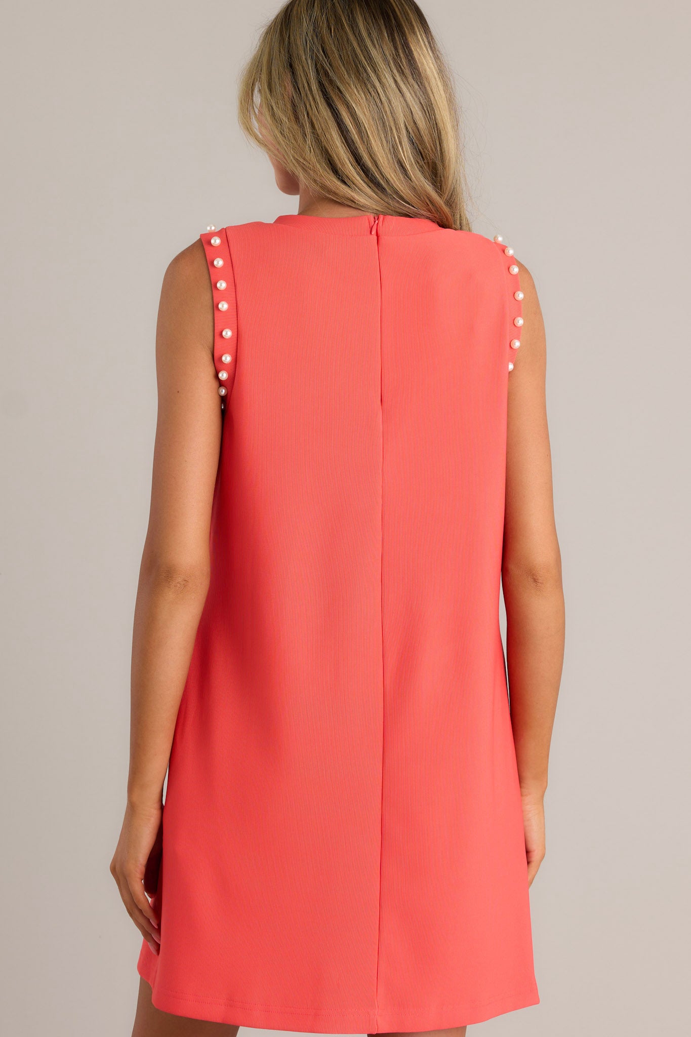 Back view of this red mini dress features a faux pearl embellished crew neckline, a discrete zipper, a ribbed texture, functional hip pockets, and a faux pearl embellished sleeveless design.