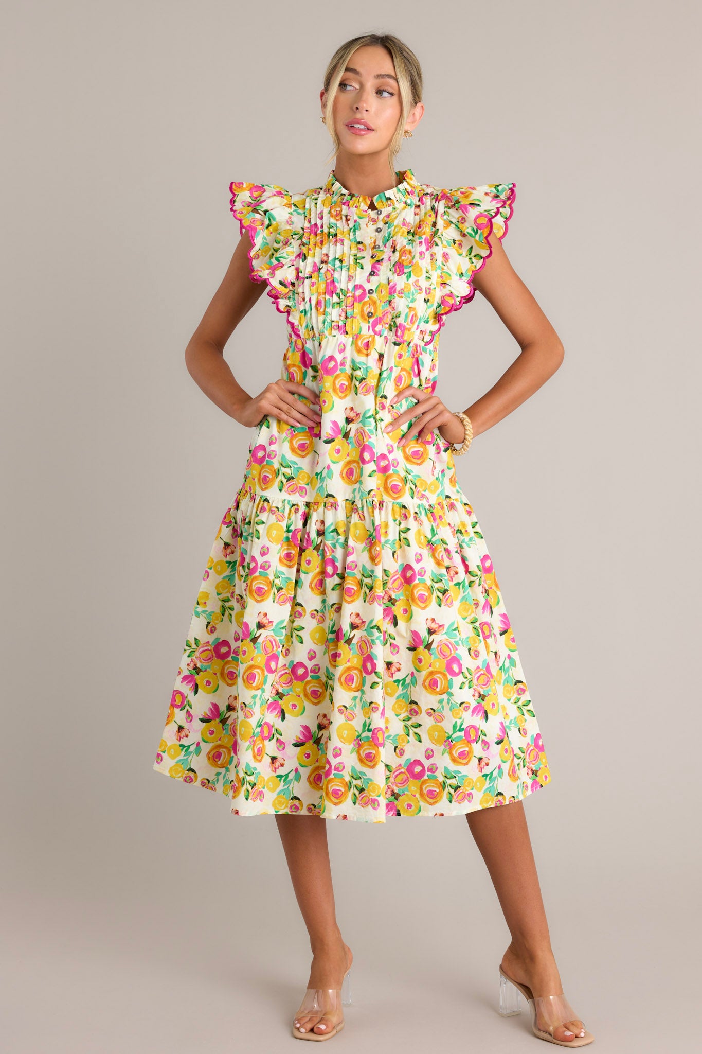 Front angled view of a yellow floral midi dress featuring a high ruffled neckline, a functional button front bodice, a pleated chest, functional hip pockets, a single tier, and short flutter sleeves