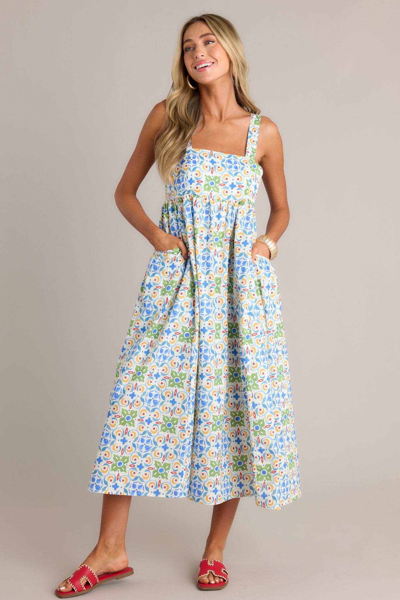 Angled full length view of a white midi dress with a square neckline, self-tie adjustable straps, a colorful pattern, functional hip pockets, and a flowing silhouette