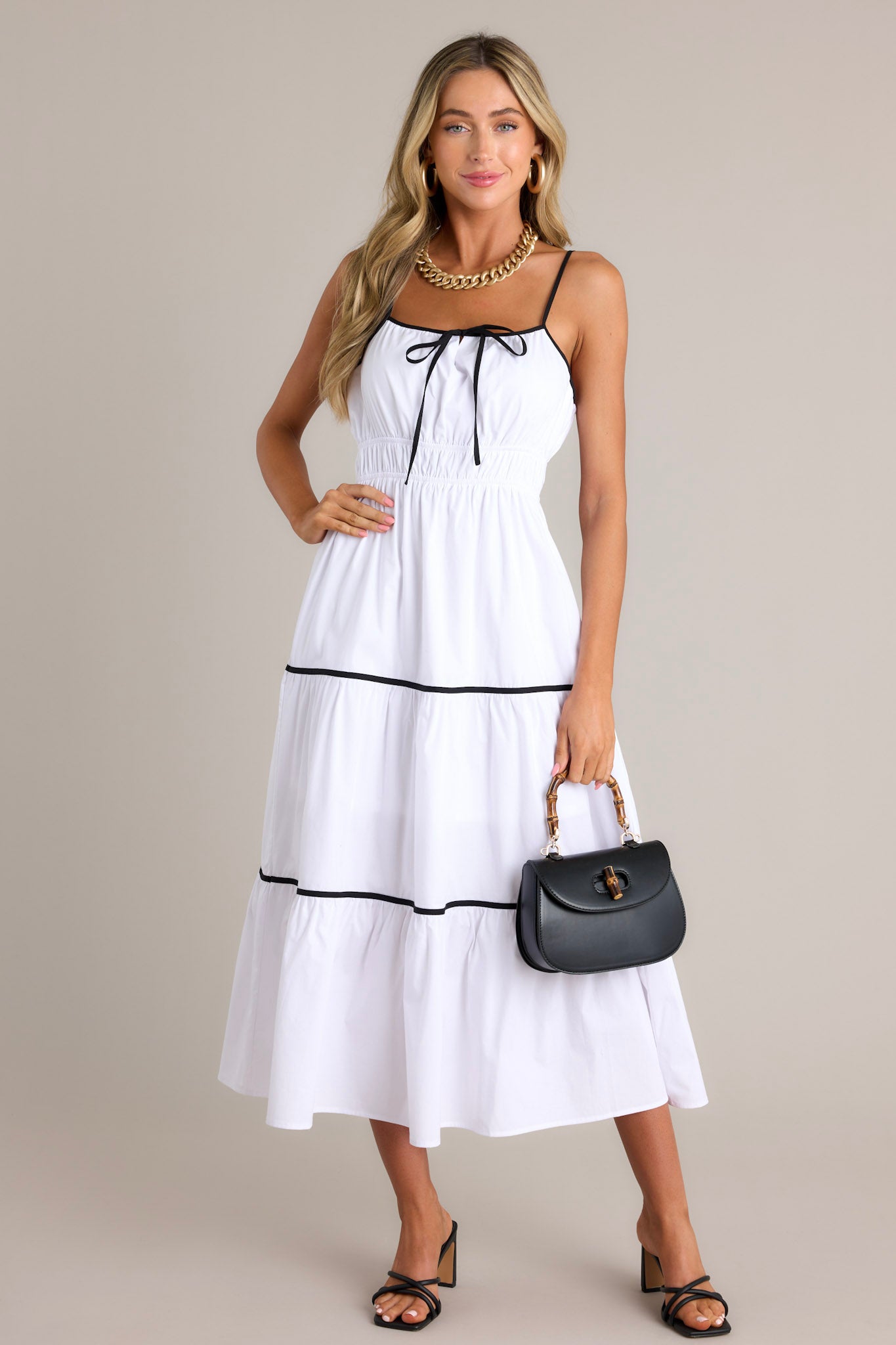 This white midi dress features a square neckline, thin adjustable straps, a self-tie bust feature, an elastic waist, contrasting tiers, and a flowing silhouette.