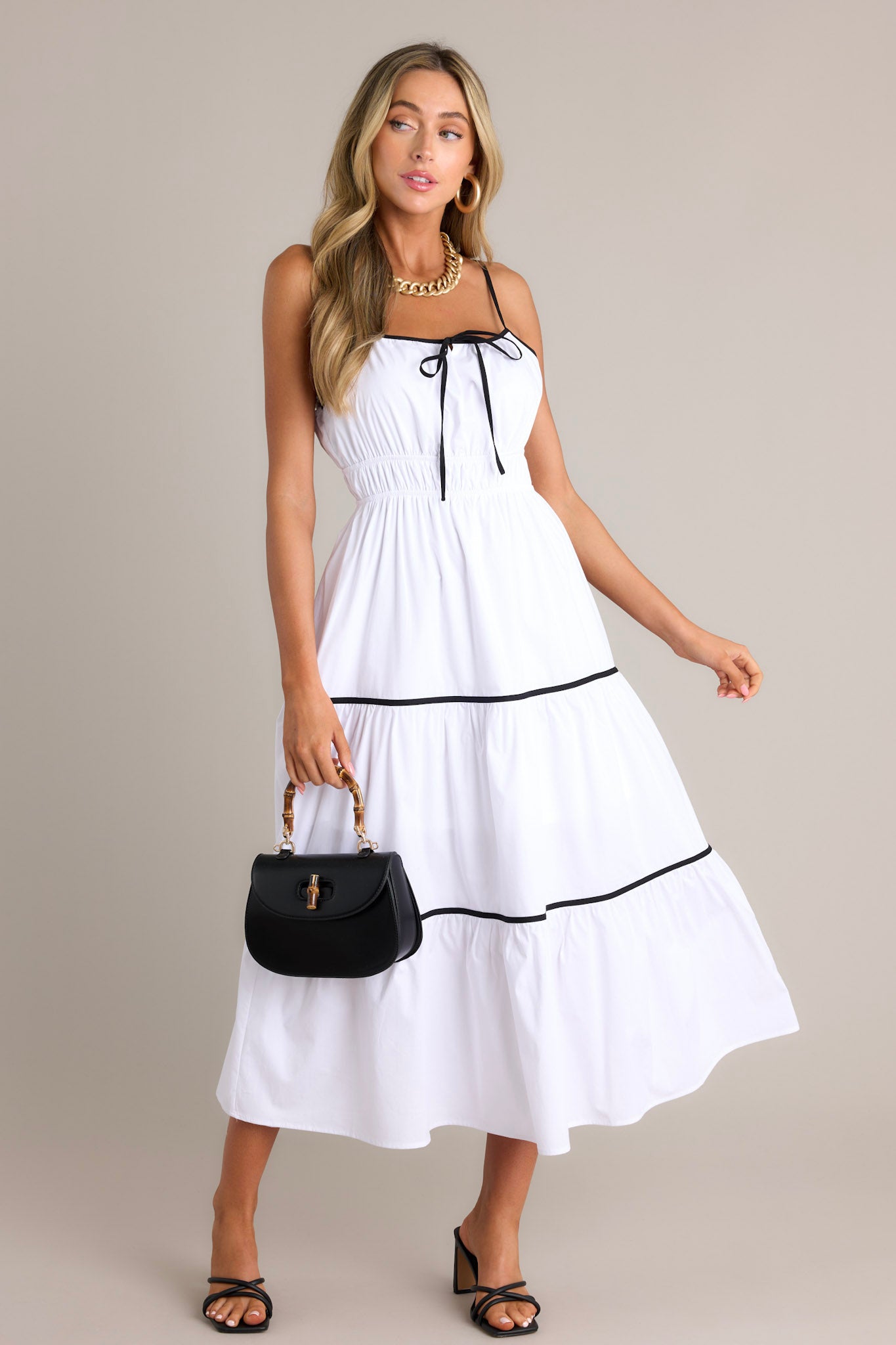 Front angled view of a white midi dress featuring a square neckline, thin adjustable straps, a self-tie bust feature, an elastic waist, contrasting tiers, and a flowing silhouette