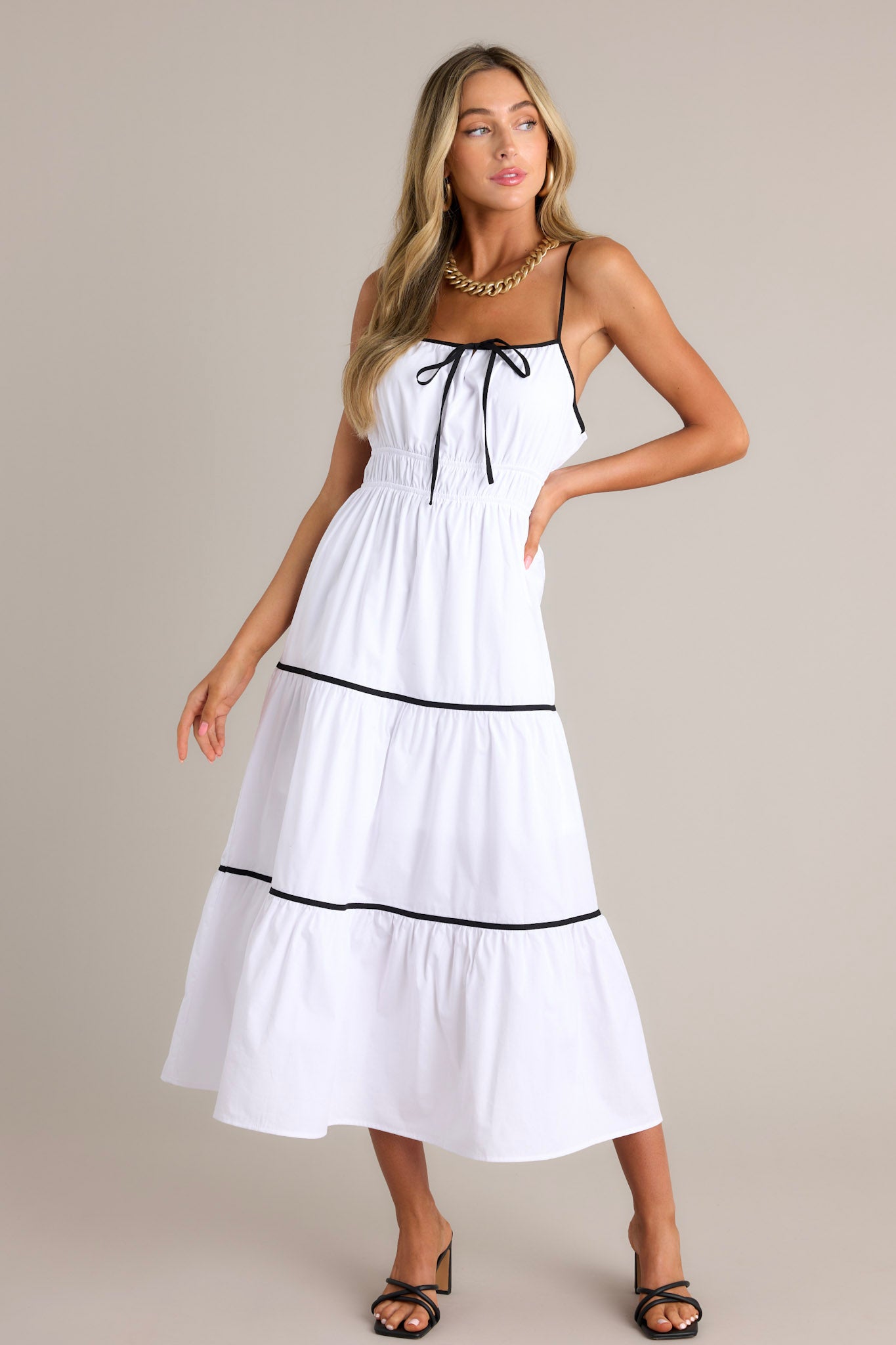 Full length view of a white midi dress with a square neckline, thin adjustable straps, a self-tie bust feature, an elastic waist, contrasting tiers, and a flowing silhouette