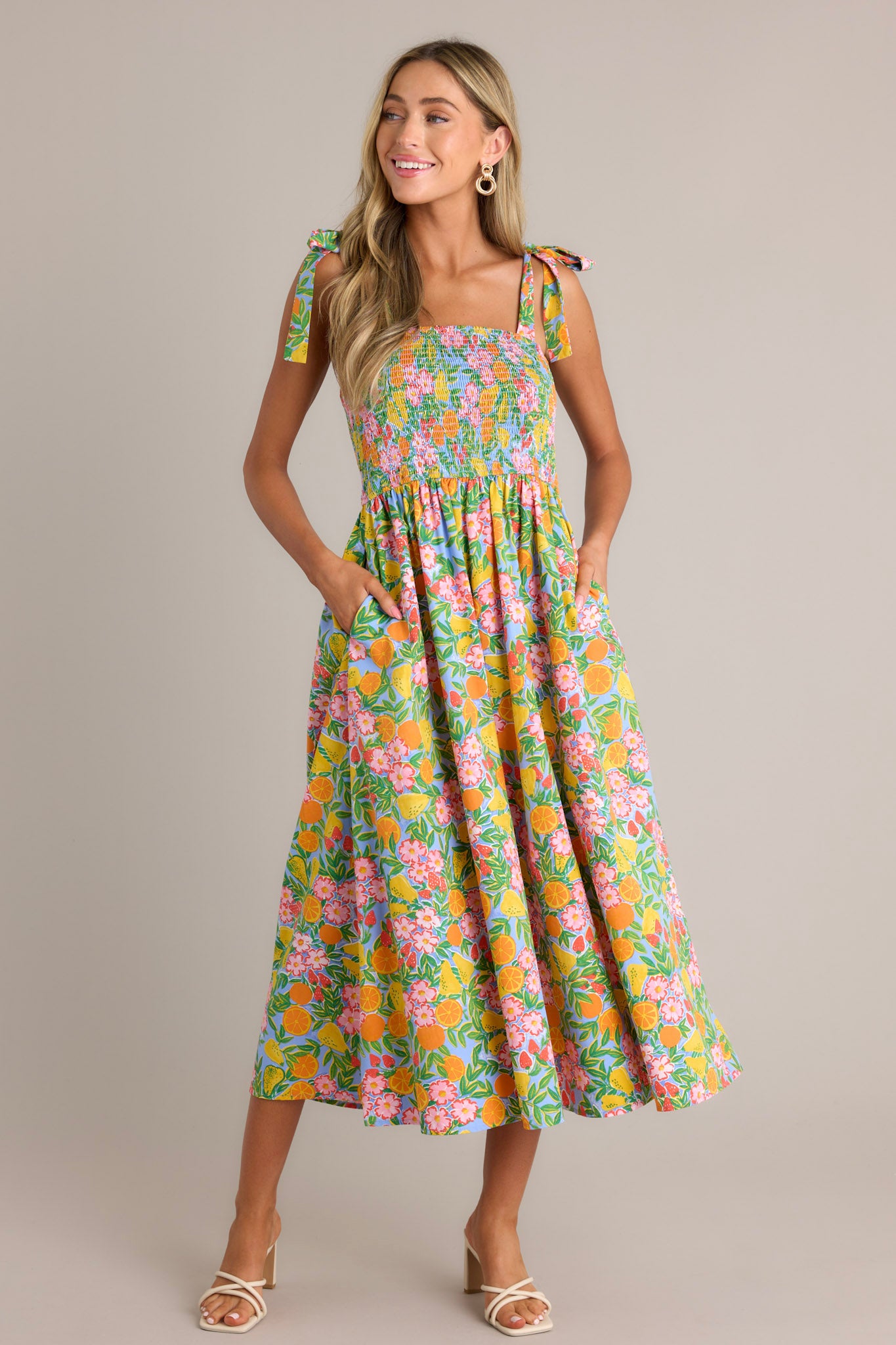 Front angled view of a green multi midi dress featuring a square neckline, self-tie shoulder straps, a fully smocked bodice, functional hip pockets, a beautiful print, and a flowing silhouette