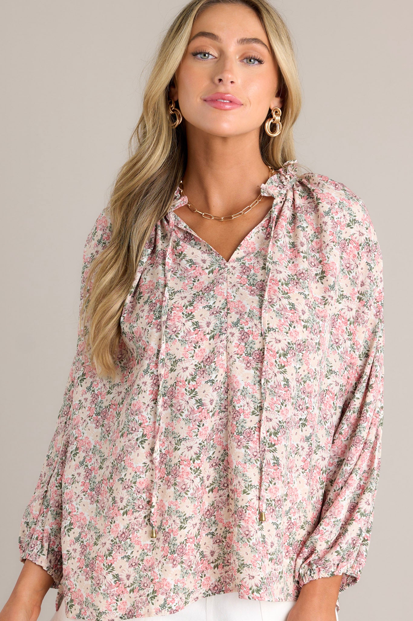 Front view of a pink floral top featuring a ruffled v-neckline, a self-tie neck feature, gathering in the back, a lightweight material, and elastic cuffed long bishop sleeves.