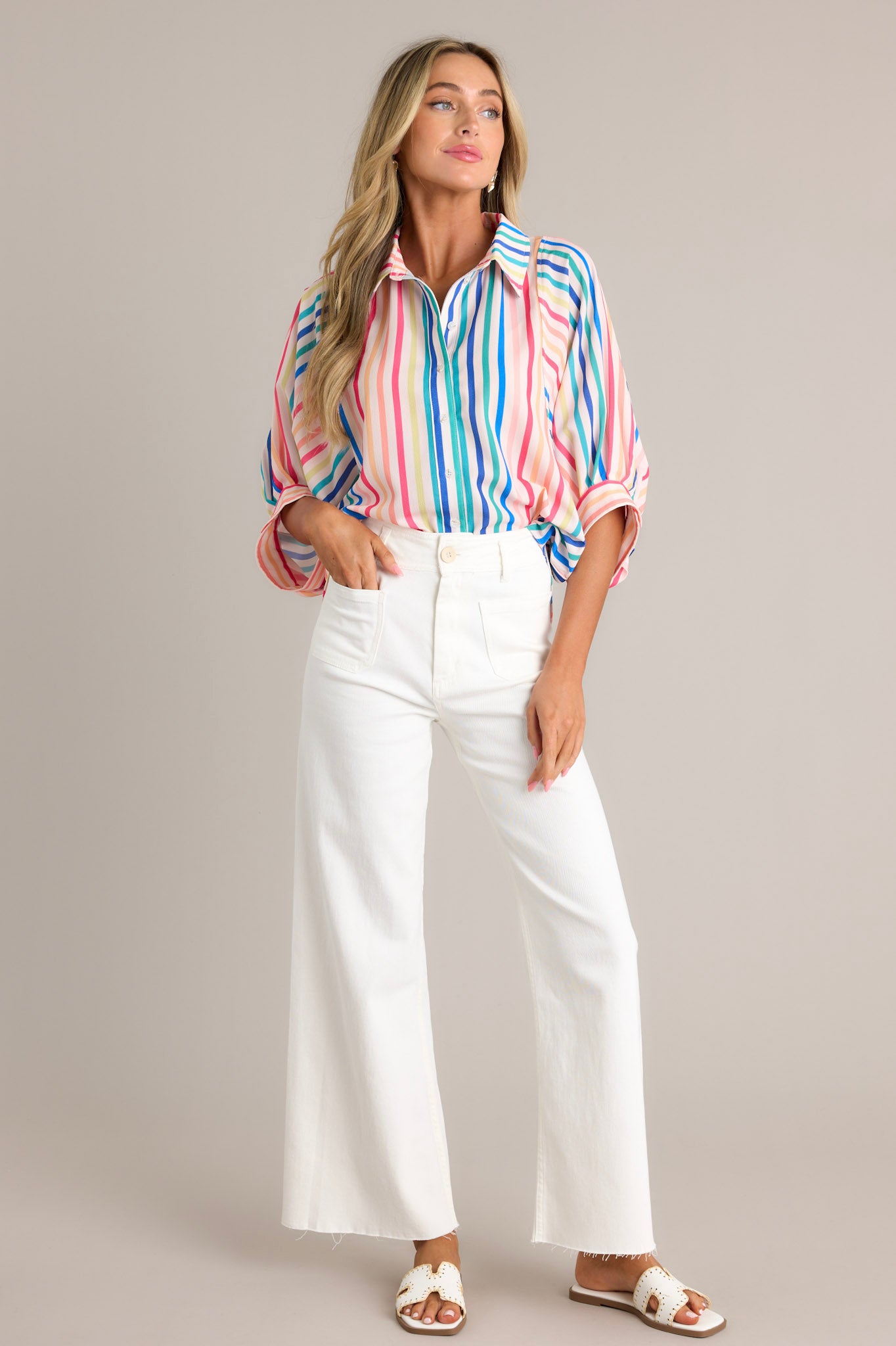 Front angled view of a rainbow stripe top featuring a collared neckline, a full button front, an oversized fit, and wide dolman button cuffed sleeves