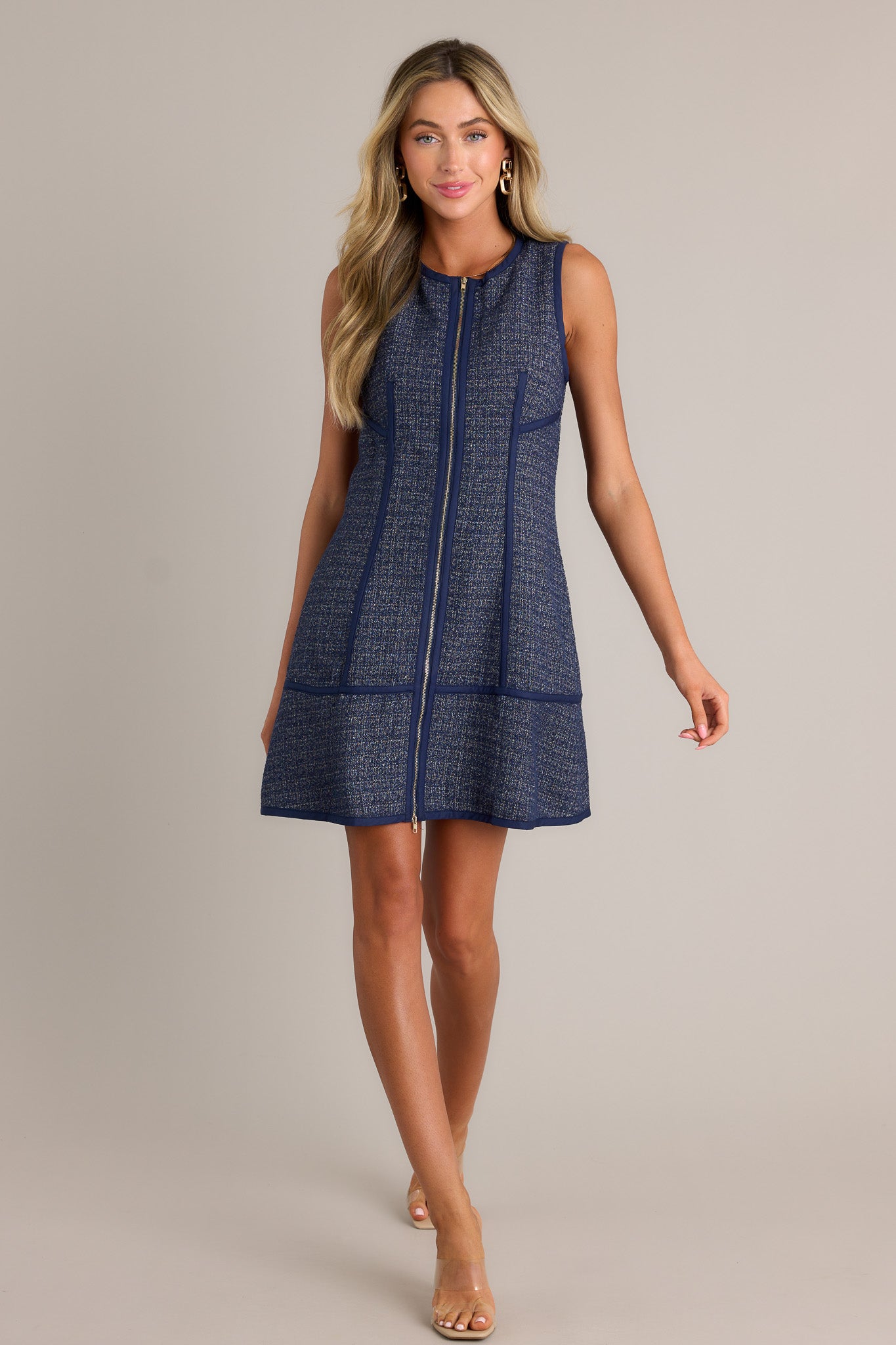 This navy mini dress features a high crew neckline, a functional zipper front, thick front seams, a tweed material, and a sleeveless design.