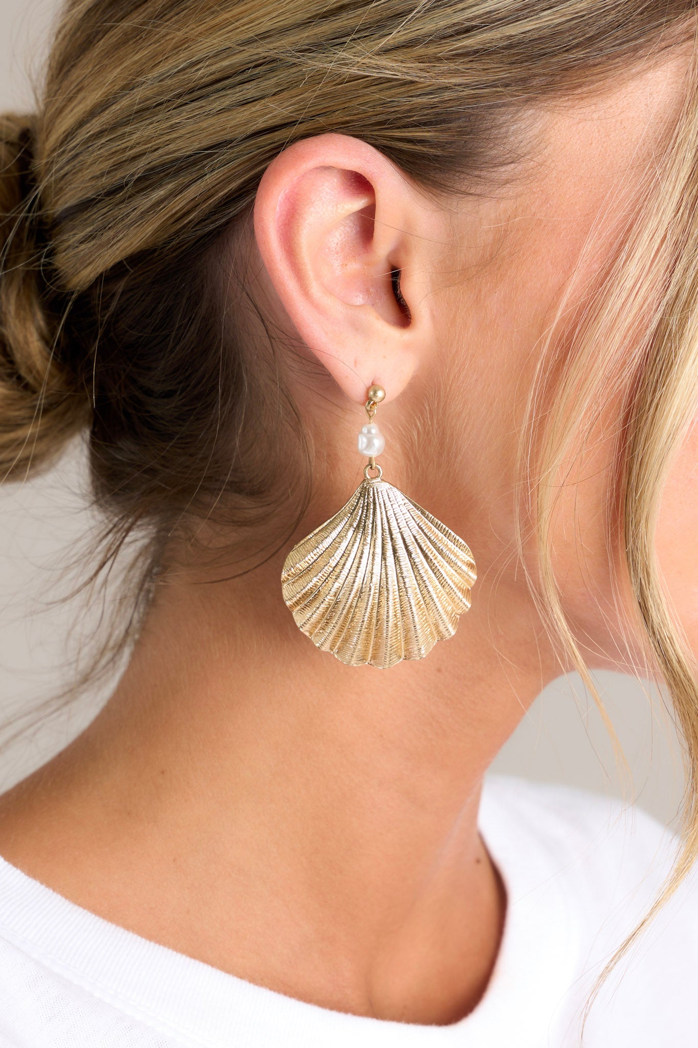 Close-up of these gold earrings that feature a gold seashell pendant and a small faux pearl detail.