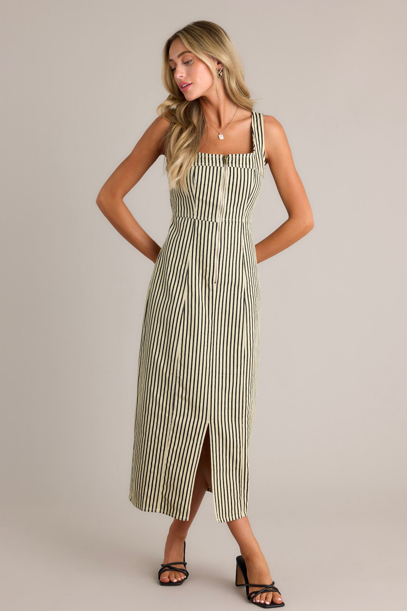 Front angled view of a black stripe maxi dress featuring a square neckline, thick straps, a functional zip front, a vertical stripe pattern, and a front slit