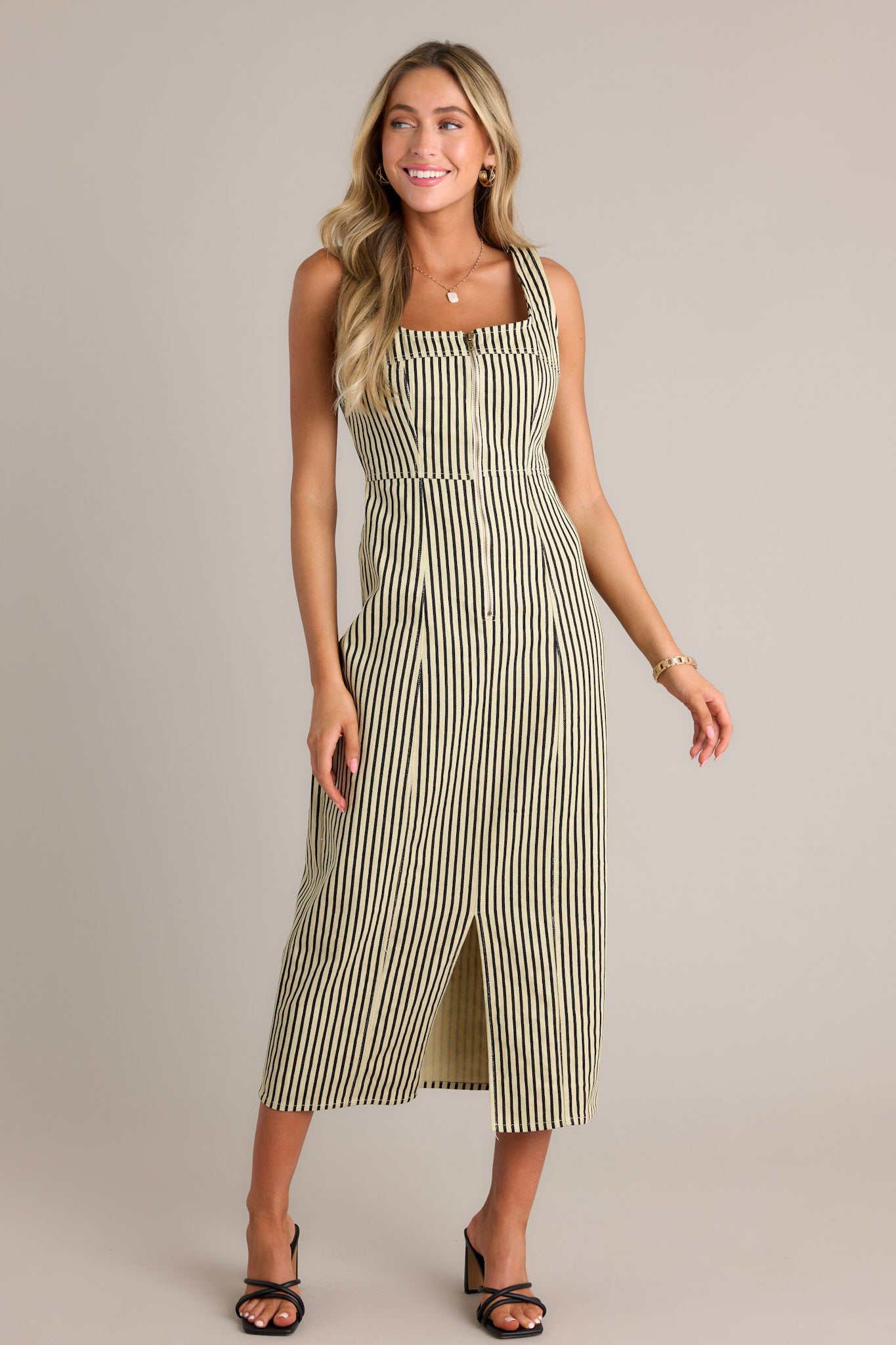 Action shot of a black stripe maxi dress displaying the fit and movement, highlighting the square neckline, thick straps, functional zip front, vertical stripe pattern, and front slit.
