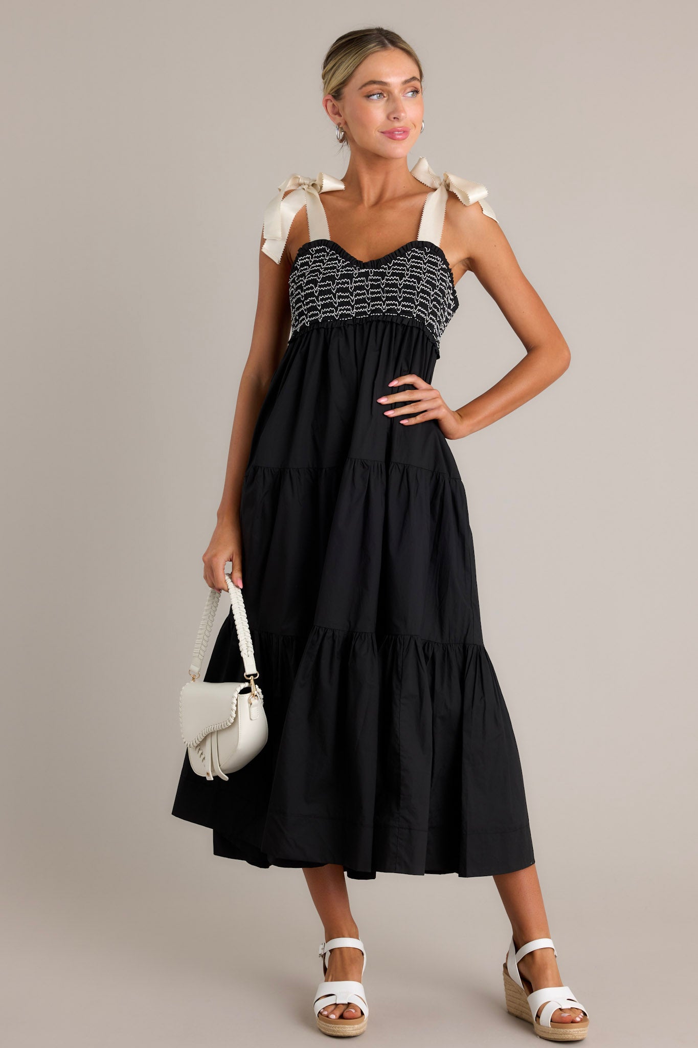 Front angled view of a black maxi dress featuring a v-neckline, thick self-tie straps, a fully smocked bodice, functional hip pockets, and a tiered design