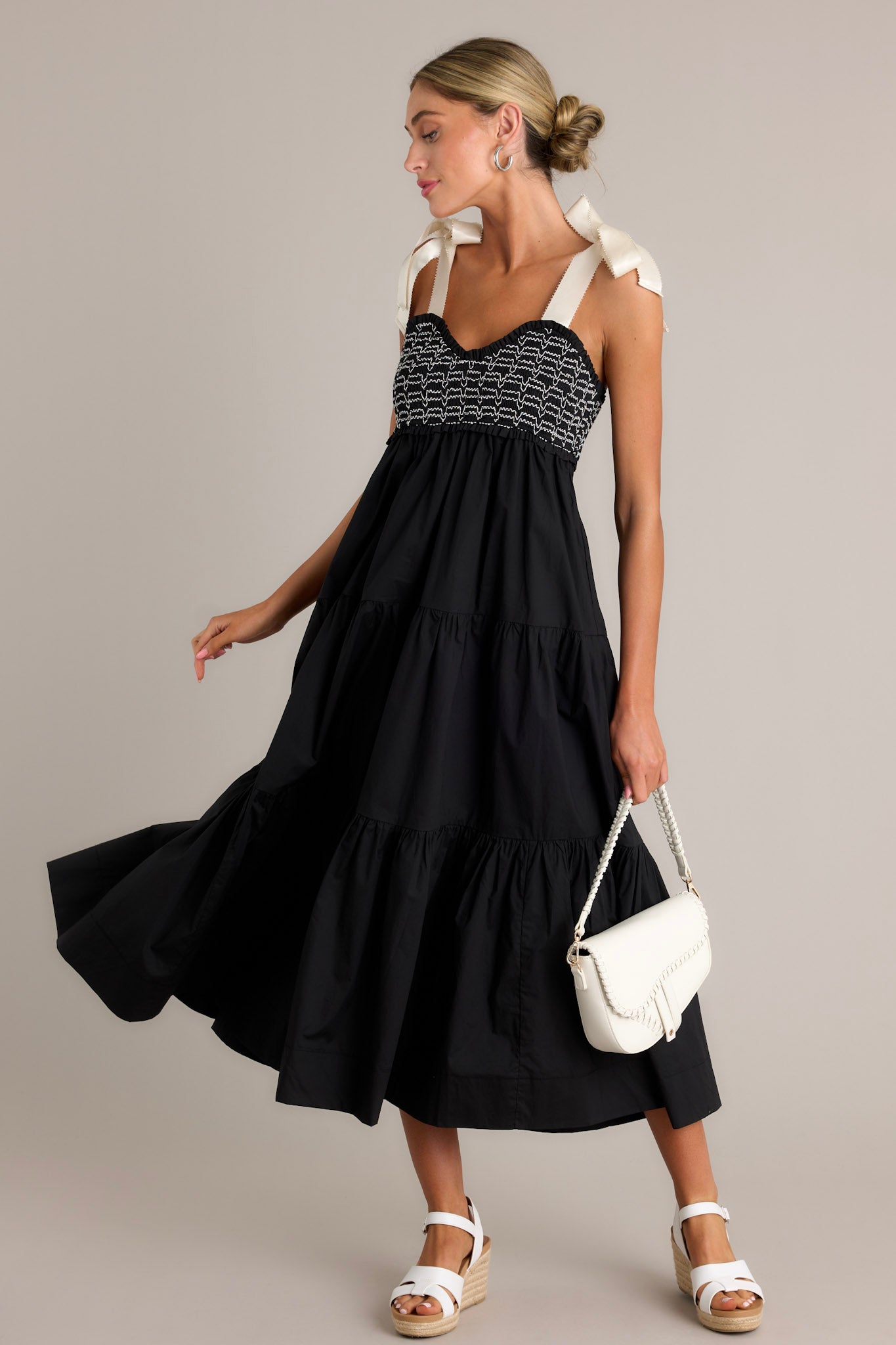 Action shot of a black maxi dress displaying the fit and movement, highlighting the v-neckline, thick self-tie straps, fully smocked bodice, functional hip pockets, and tiered design.