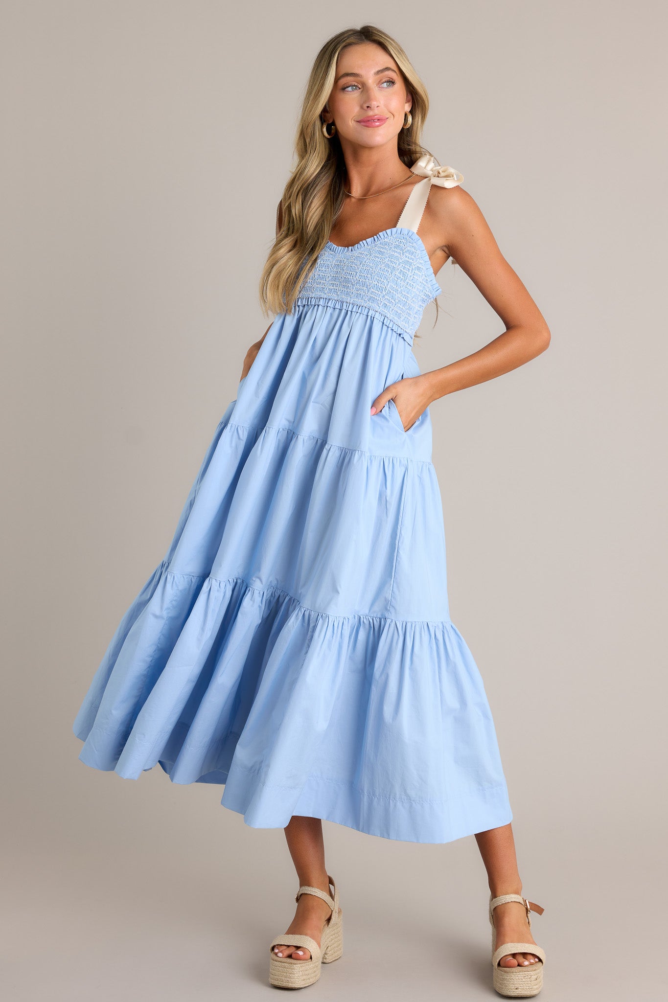 Full length view of a light blue maxi dress with a v-neckline, thick self-tie straps, a fully smocked bodice, functional hip pockets, and a tiered design