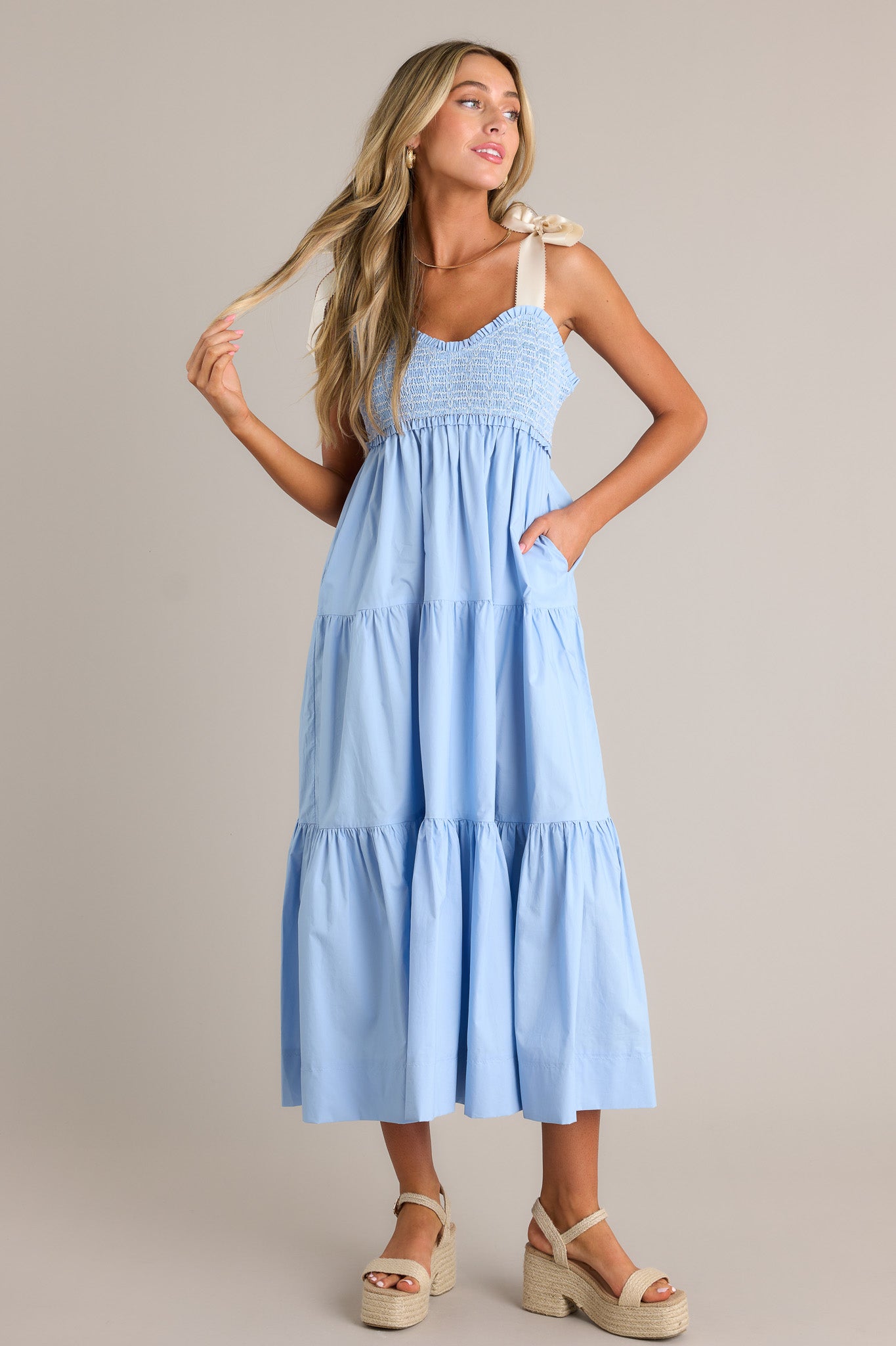 This light blue maxi dress features a v-neckline, thick self-tie straps, a fully smocked bodice, functional hip pockets, and a tiered design.