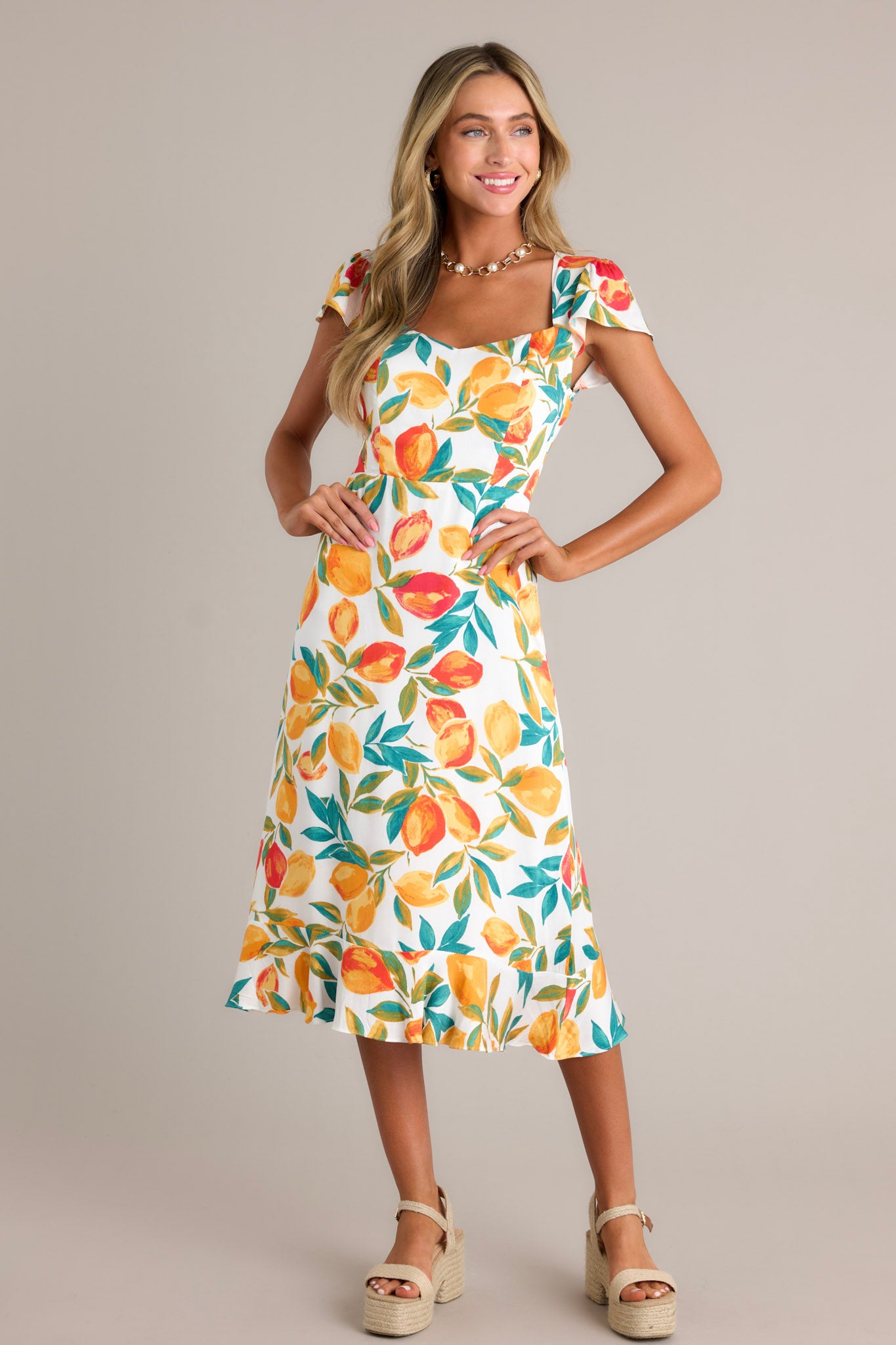 Front angled view of an ivory dress featuring a v-neckline, a smocked back insert, a zipper, a citrus print, a ruffled hemline, and short flutter sleeves