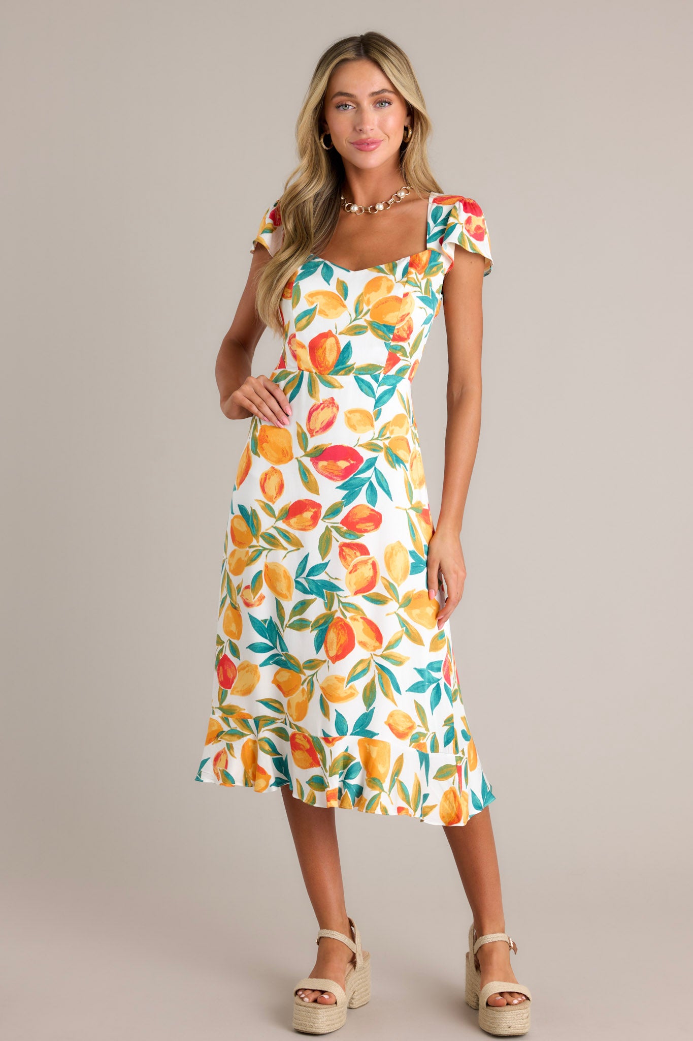 Full length view of an ivory dress with a v-neckline, a smocked back insert, a zipper, a citrus print, a ruffled hemline, and short flutter sleeves