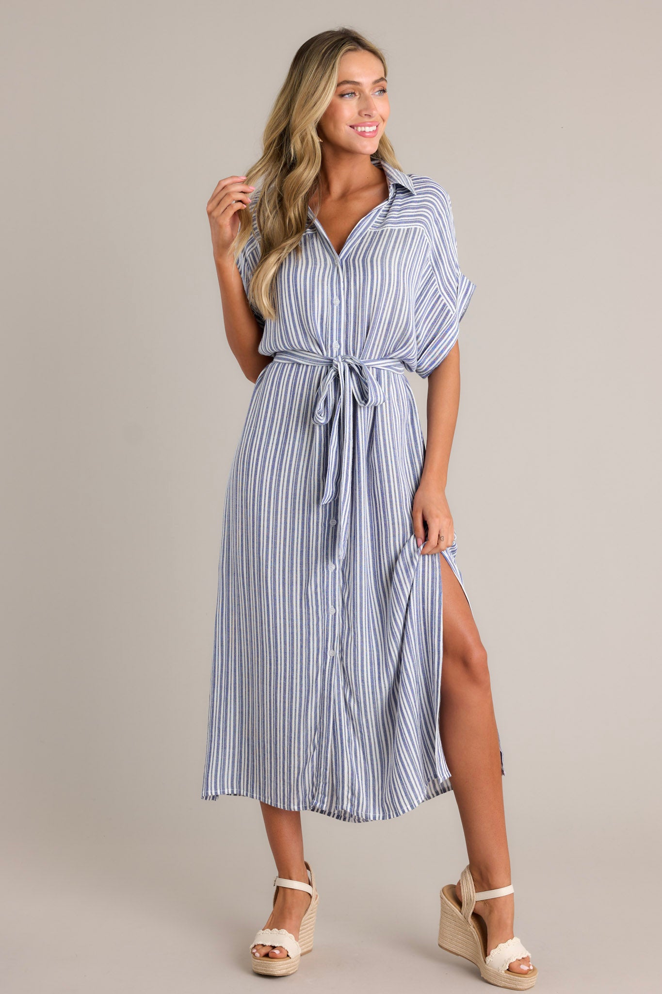Full length view of a blue stripe midi dress with a collared v-neckline, a functional button front, a self-tie drawstring belt, functional pockets, side slits, and a flowing silhouette