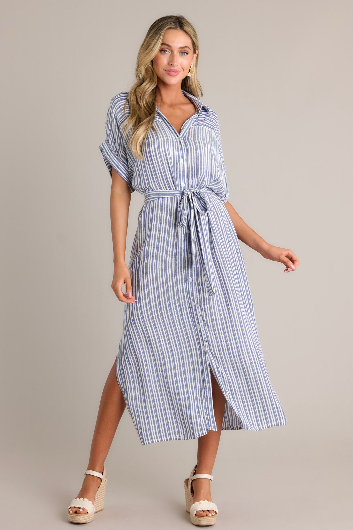 Action shot of a blue stripe midi dress displaying the fit and movement, highlighting the collared v-neckline, functional button front, self-tie drawstring belt, functional pockets, side slits, and flowing silhouette.