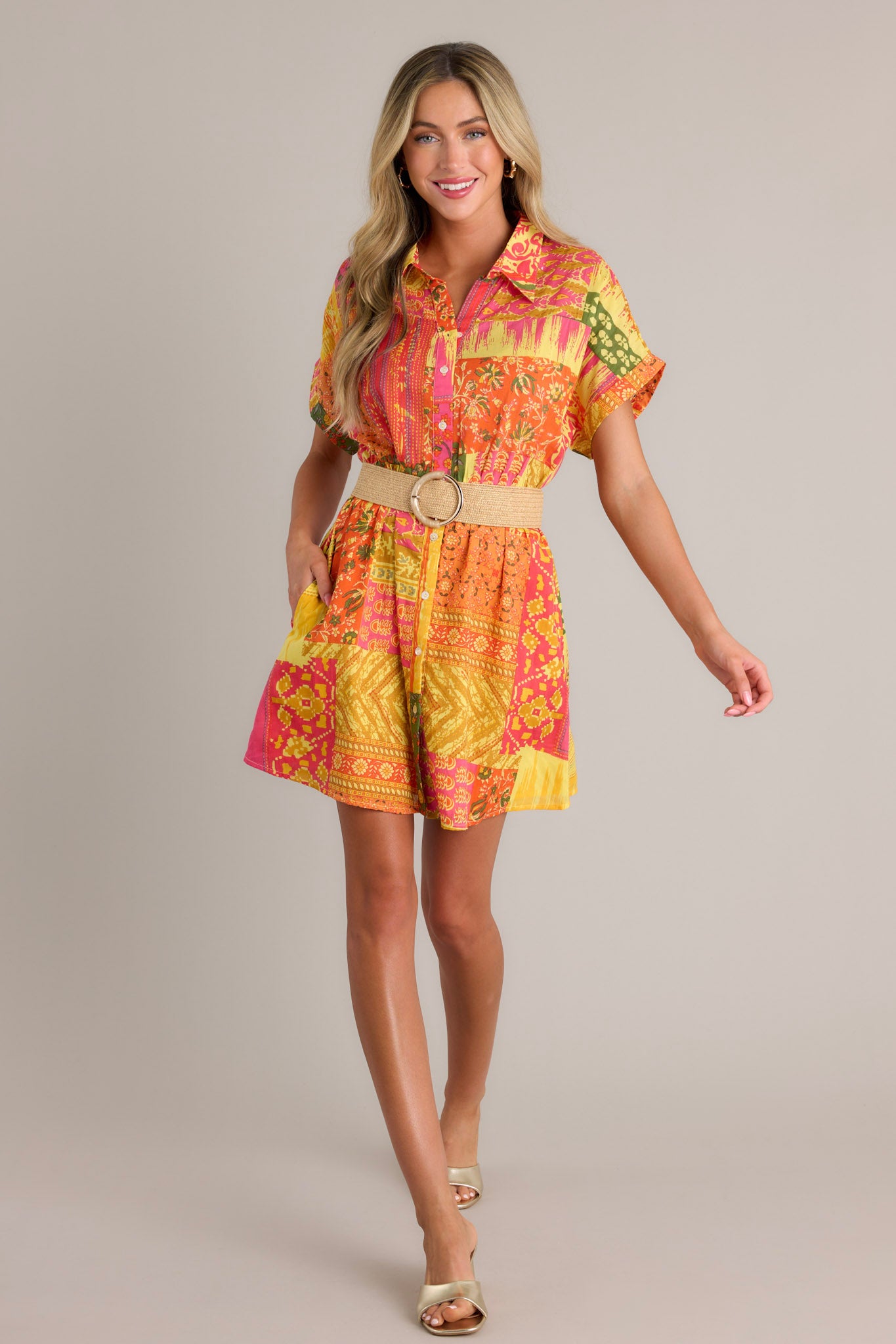 Full length view of an orange mini dress with a collared neckline, a full button front, a unique patchwork design, an elastic waistband, a self-tie waist belt, functional hip pockets, and cuffed short sleeves