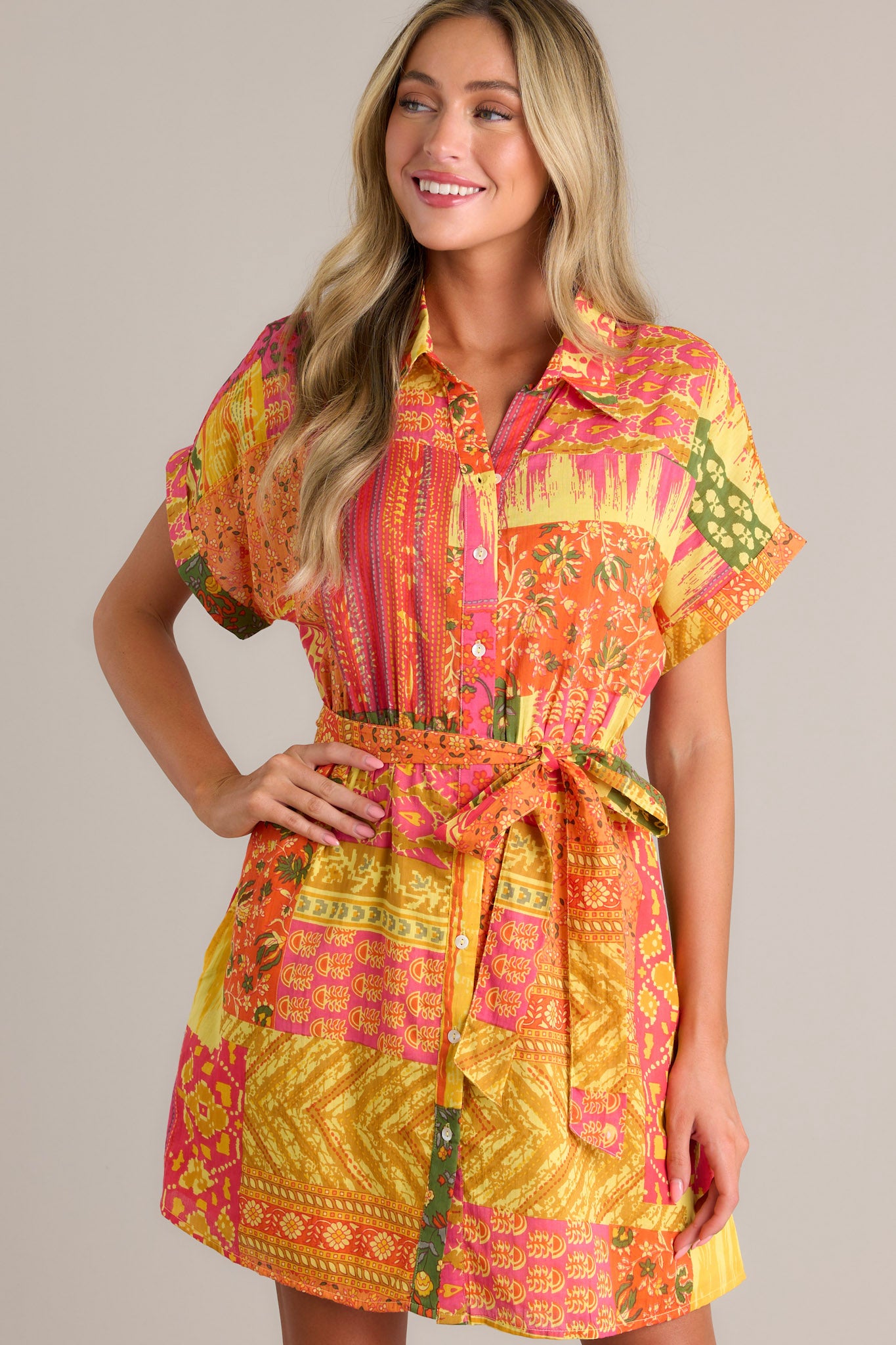 Front angled view of an orange mini dress featuring a collared neckline, a full button front, a unique patchwork design, an elastic waistband, a self-tie waist belt, functional hip pockets, and cuffed short sleeves