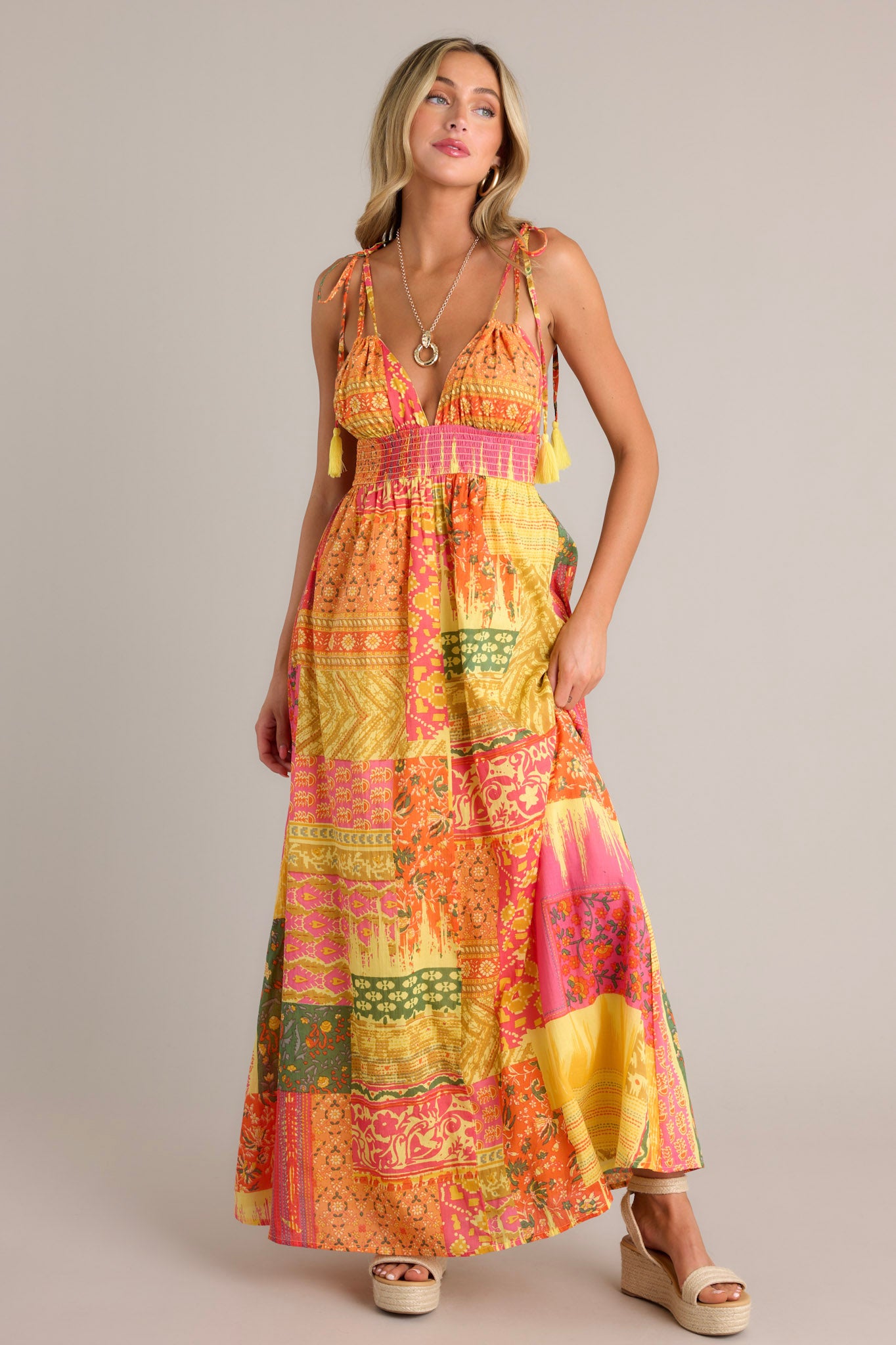 Front angled view of an orange maxi dress featuring a v-neckline, adjustable self-tie straps, a low back, a fully smocked waistband, a unique patchwork design, and a flowing silhouette