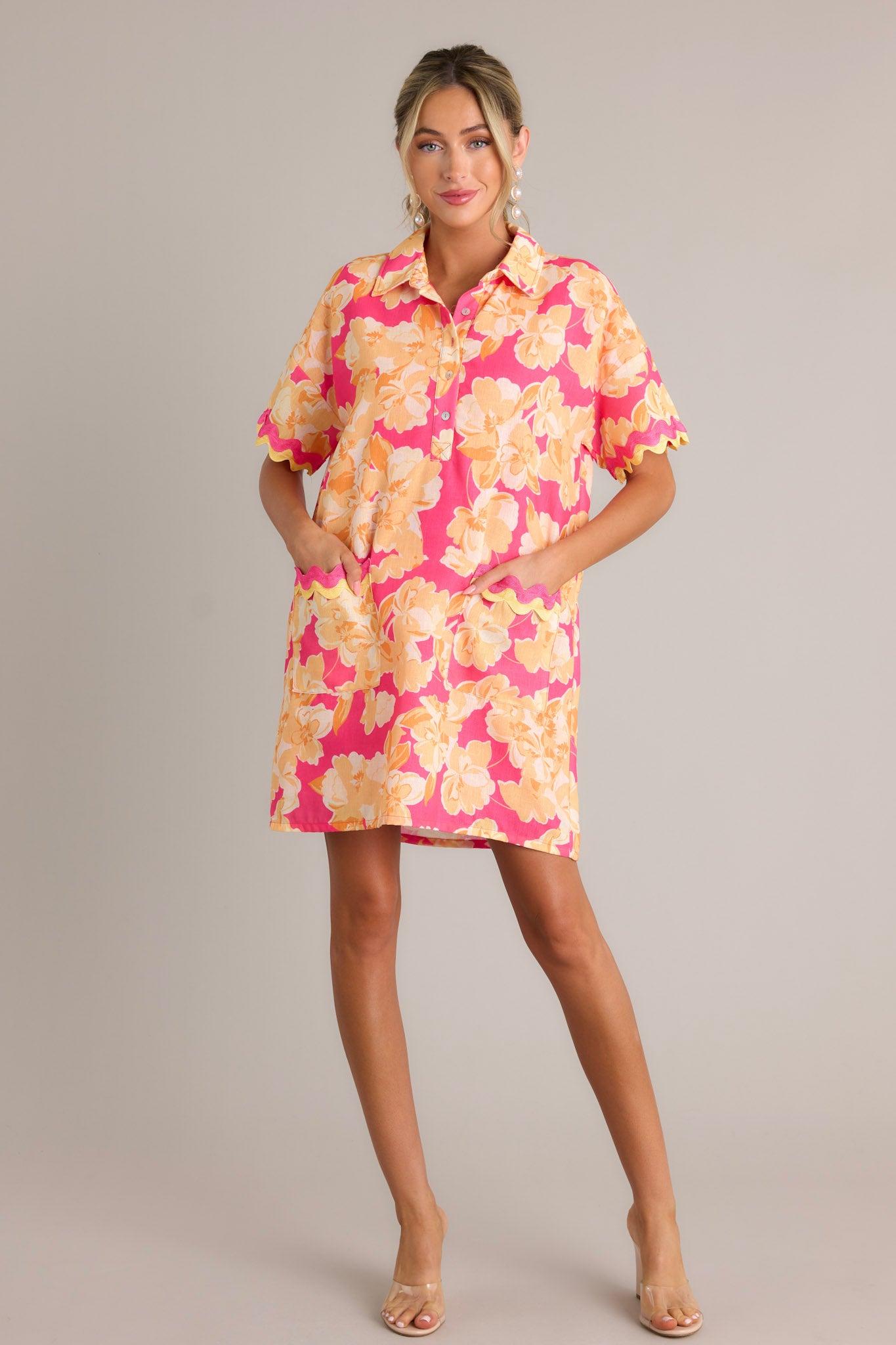 Full length view of a pink floral mini dress with a collared neckline, a functional half button front, functional front pockets with ricrac detailing, short sleeves with ricrac trim, and a relaxed fit