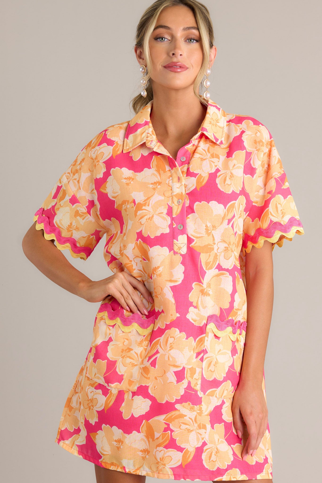 Front view of a pink floral mini dress featuring a collared neckline, a functional half button front, functional front pockets with ricrac detailing, short sleeves with ricrac trim, and a relaxed fit.