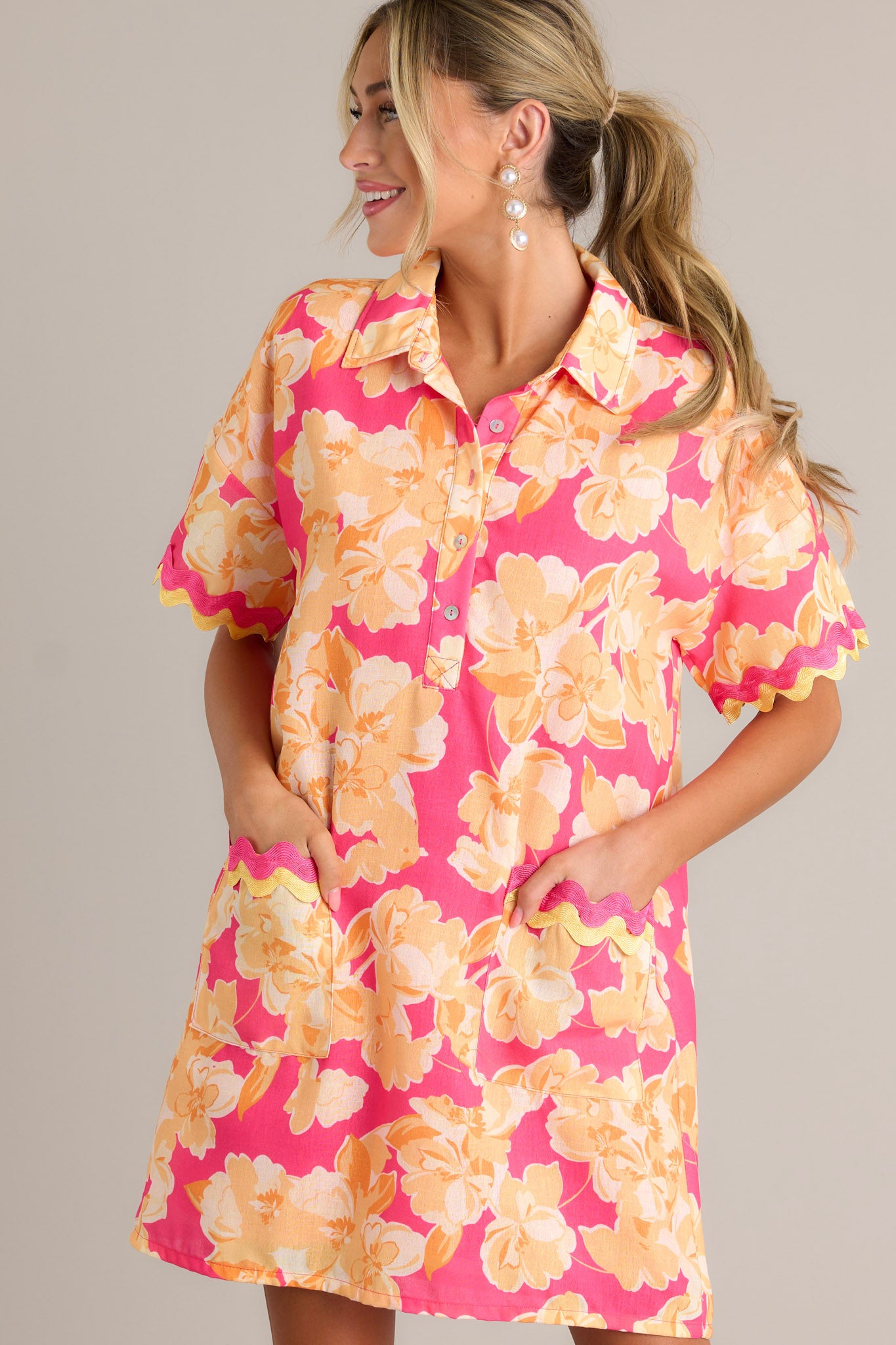 Front angled view of a pink floral mini dress featuring a collared neckline, a functional half button front, functional front pockets with ricrac detailing, short sleeves with ricrac trim, and a relaxed fit