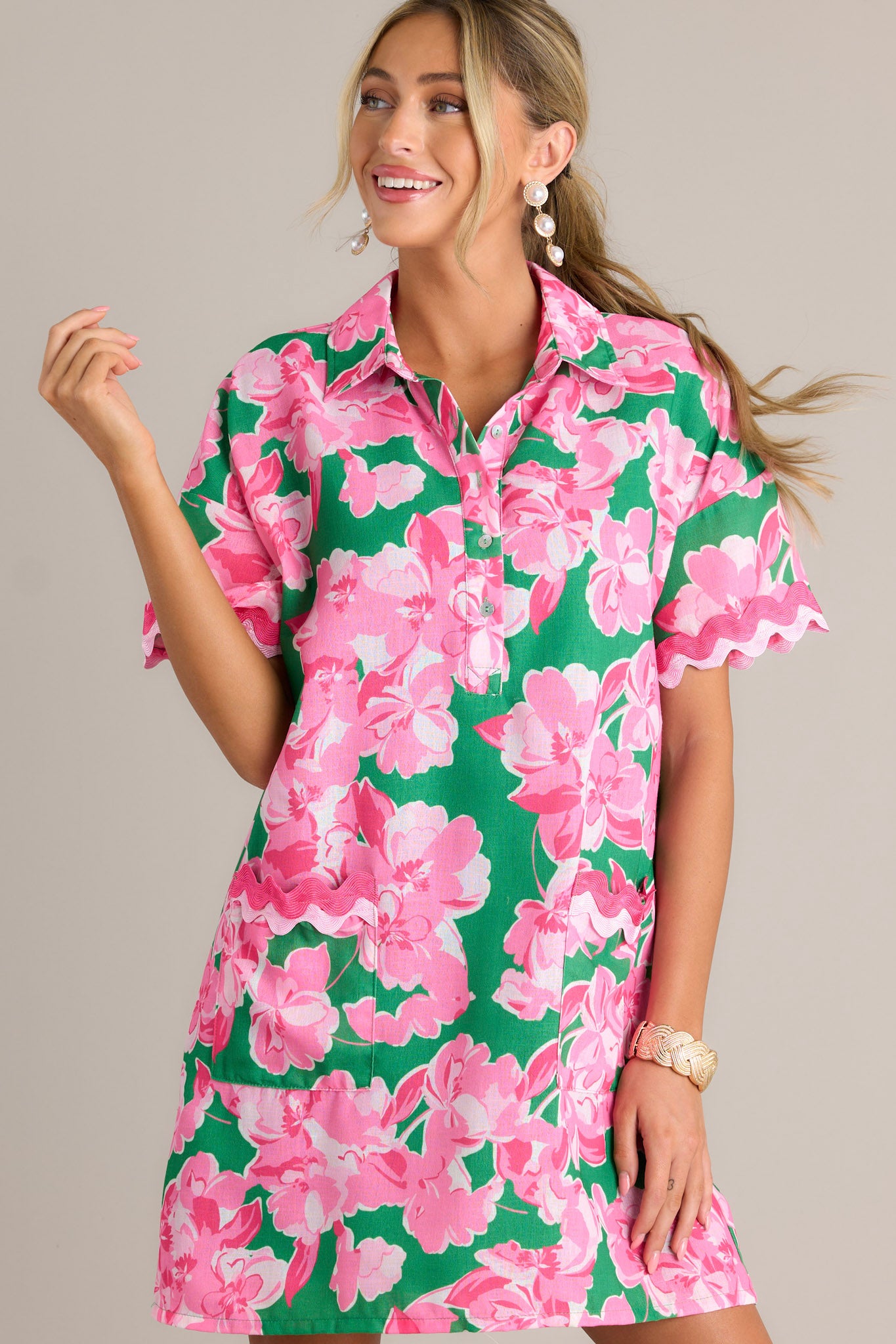 Front angled view of a green floral mini dress featuring a collared neckline, a functional half button front, functional front pockets with ricrac detailing, short sleeves with ricrac trim, and a relaxed fit