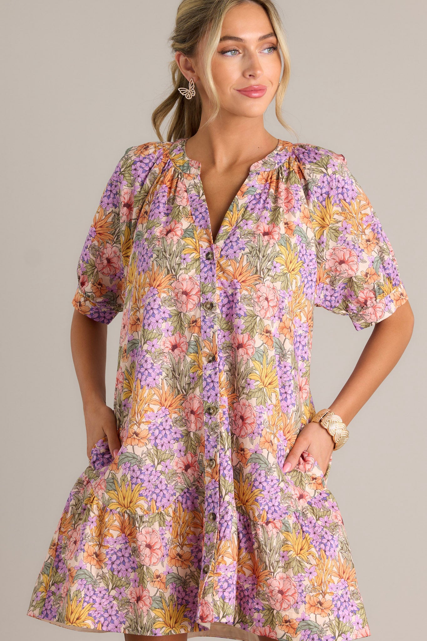 Front angled view of a lavender mini dress featuring a v-neckline, gathering in the chest, a functional button front, functional hip pockets, a scalloped tier, and button cuffed half sleeves