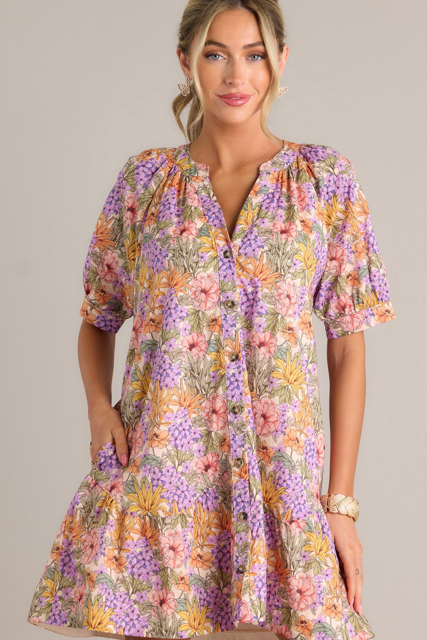 Front view of a lavender mini dress featuring a v-neckline, gathering in the chest, a functional button front, functional hip pockets, a scalloped tier, and button cuffed half sleeves.