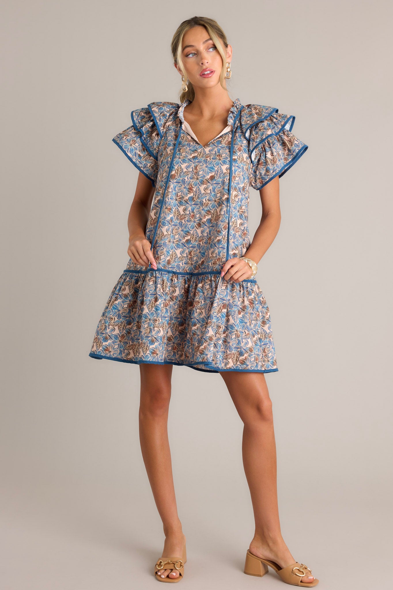 Front angled view of a blue floral mini dress featuring a ruffled v-neckline with a self-tie feature, a relaxed fit, functional hip pockets, a single tier, and ruffled short sleeves