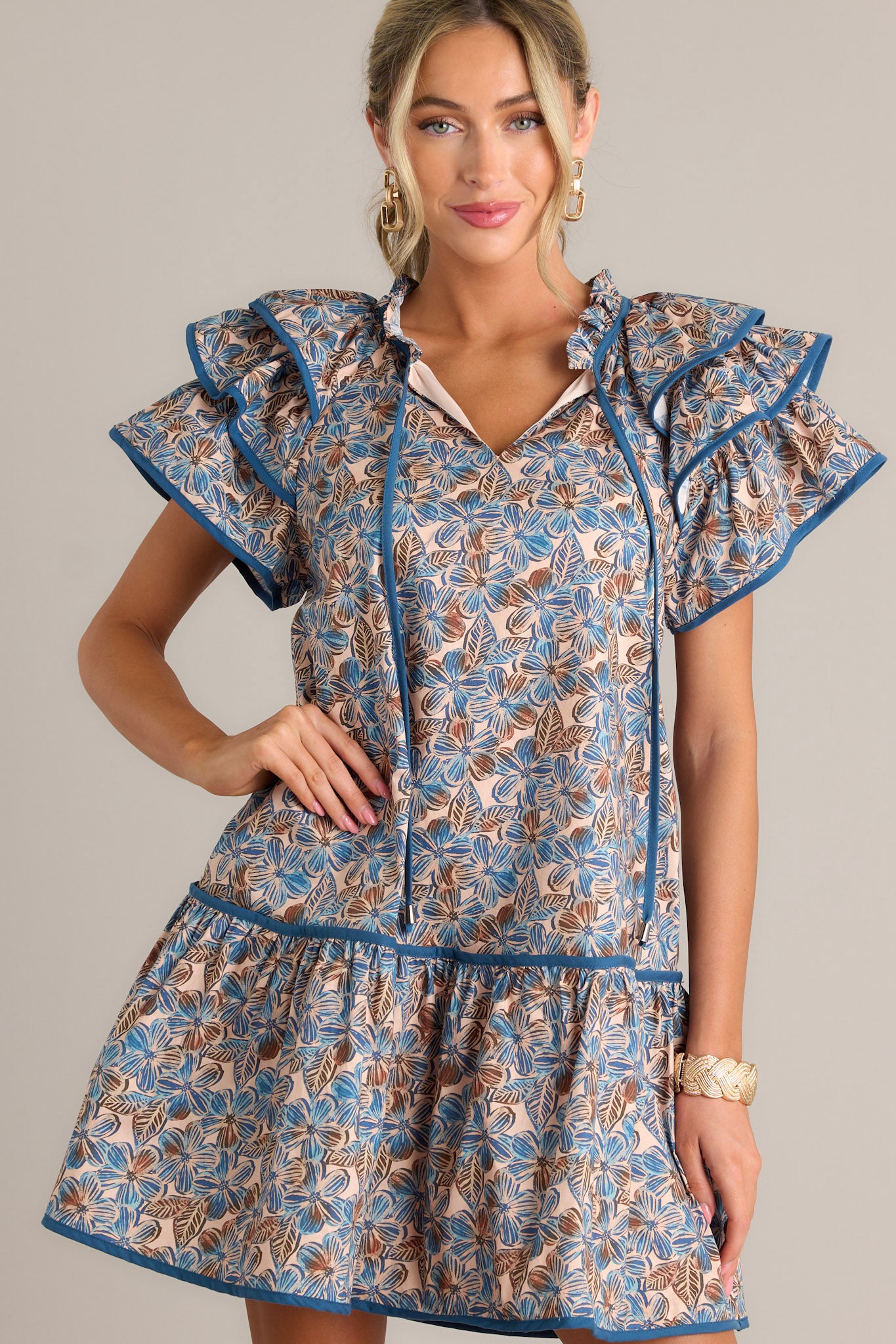 Front view of a blue floral mini dress featuring a ruffled v-neckline with a self-tie feature, a relaxed fit, functional hip pockets, a single tier, and ruffled short sleeves.