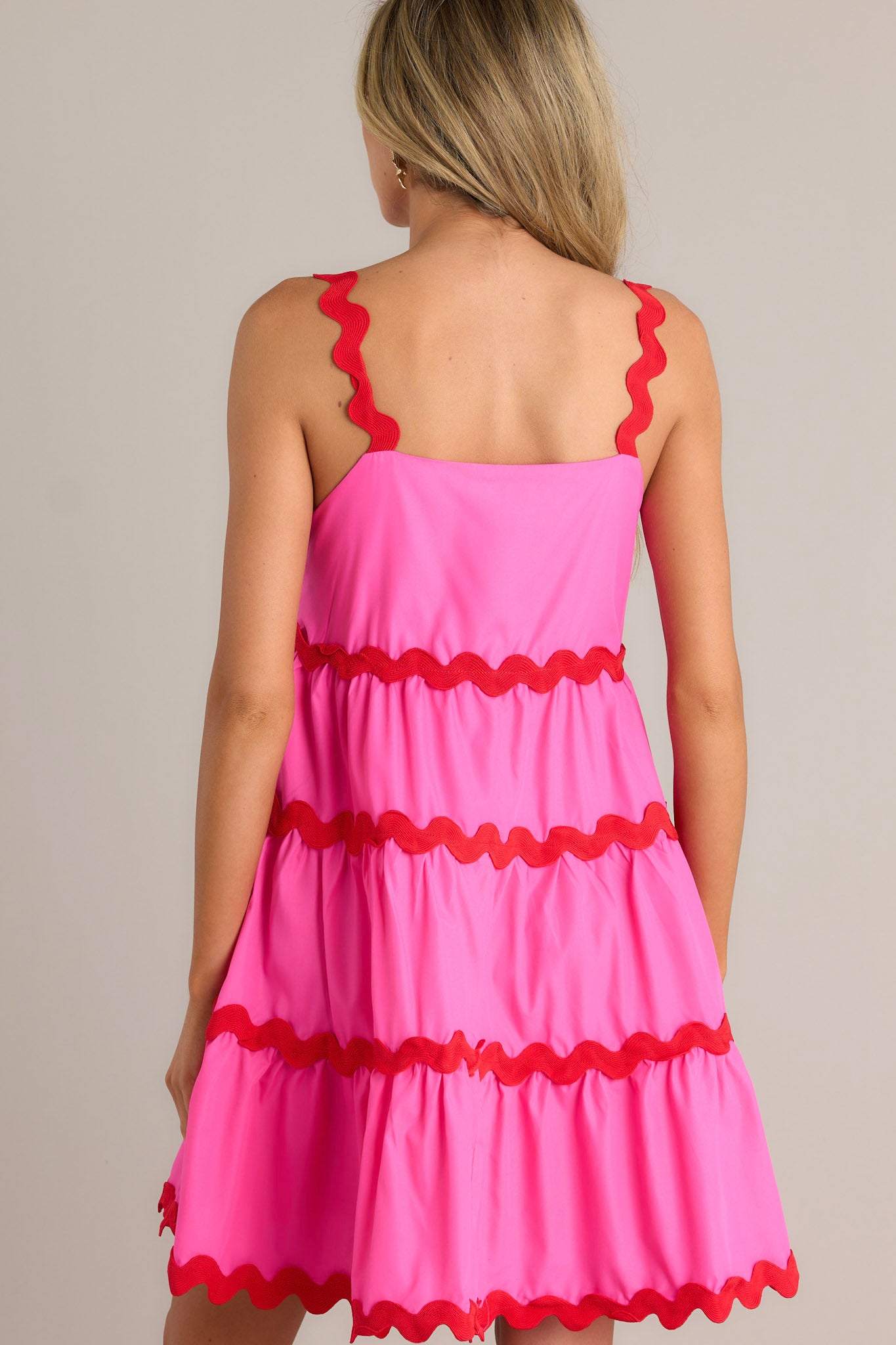 Back view of a hot pink mini dress highlighting the ricrac straps, ricrac tiers, and overall fit.