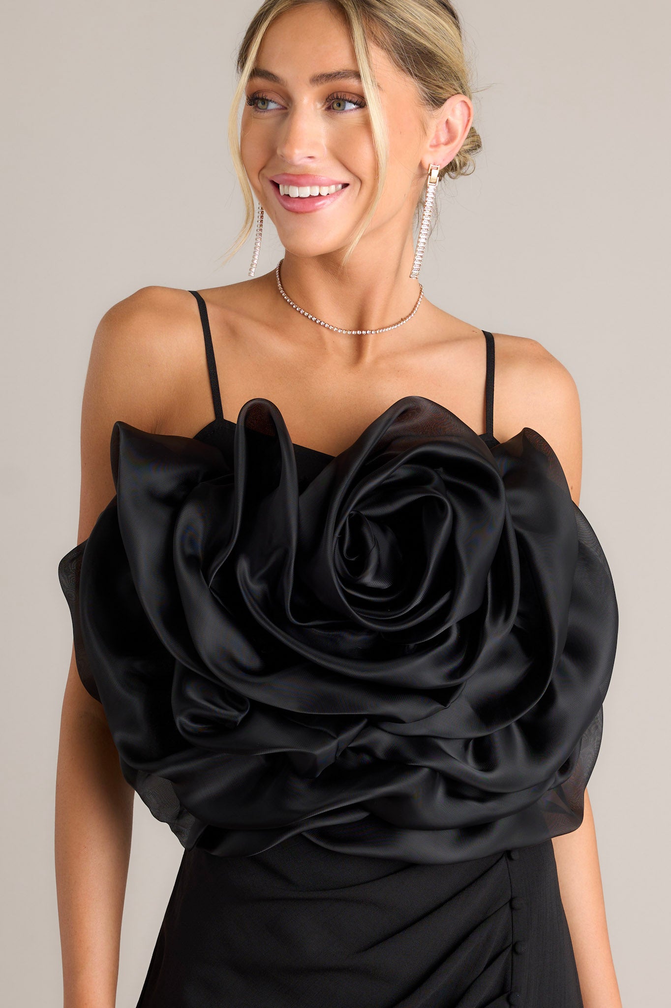 Front angled view of a black crop top featuring a square neckline, thin and stretchy adjustable straps, a fully smocked back, a large 3-D flower, and a cropped hemline