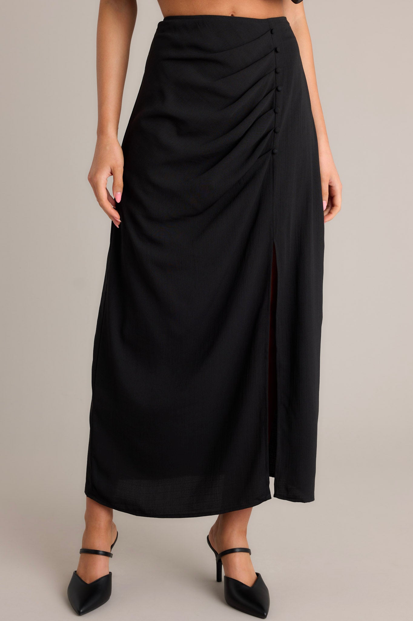 Front view of this black midi skirt featuring a high waisted design, pleated detailing on the hip, faux buttons, a discrete side zipper, and a side slit.